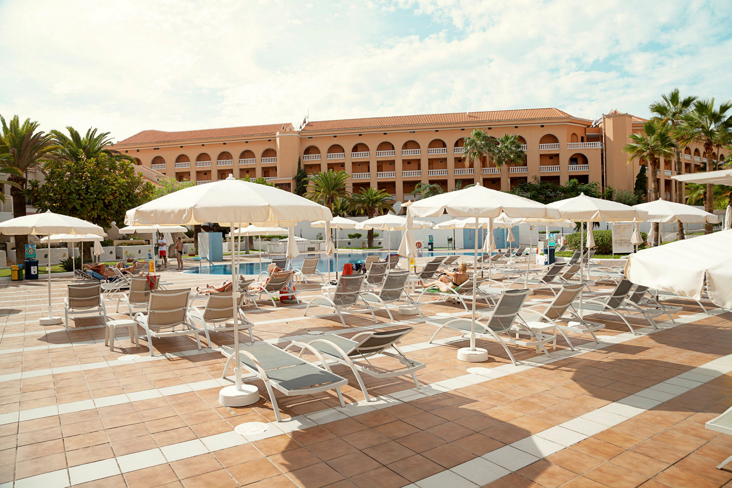 Solstolar under parasoll på en terrass nära en pool vid en hotellbyggnad. (AI-genererad text)