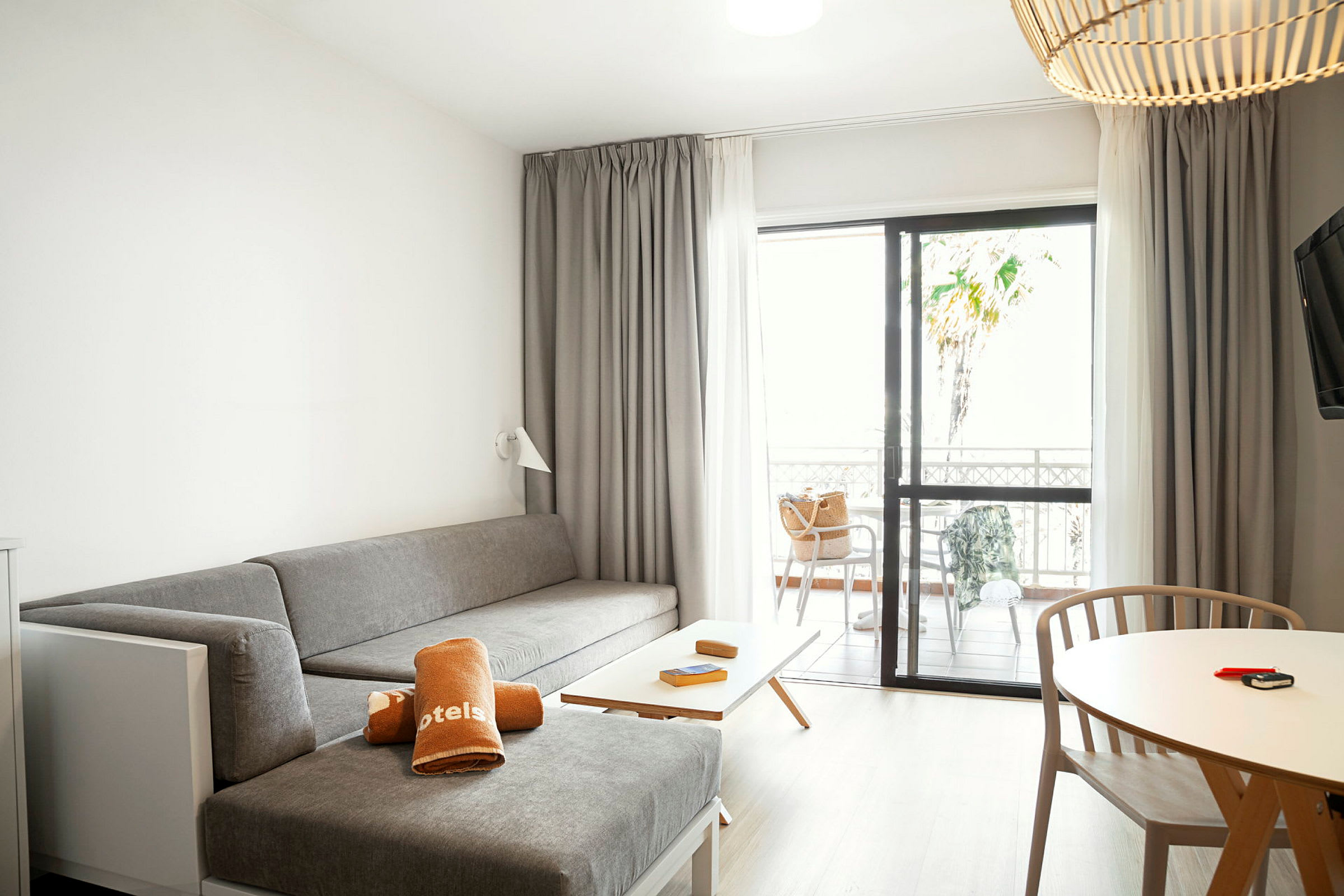 Ett hotellrum med ett grått sittområde, ett bord med trästolar och en uteplats genom glasdörrar. (AI-genererad text)