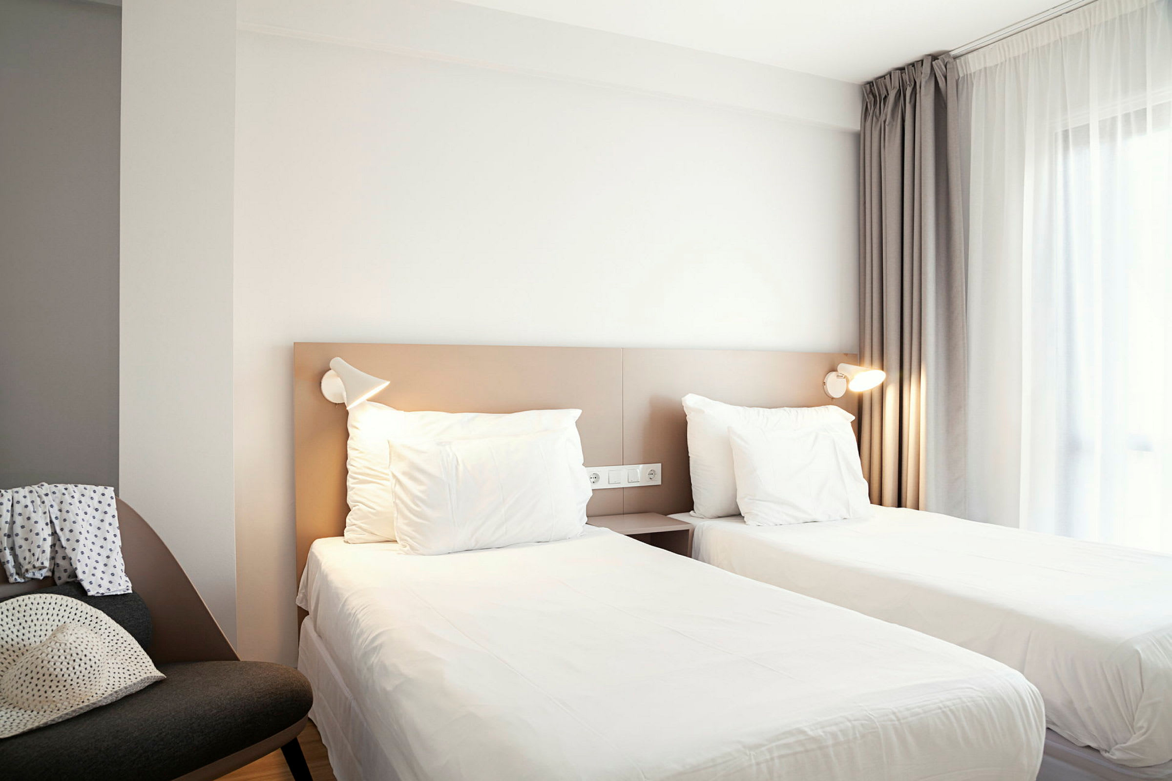 Ett hotellrum med två enkelsängar, ett fönster med gardiner och en stol på sidan. (AI-genererad text)