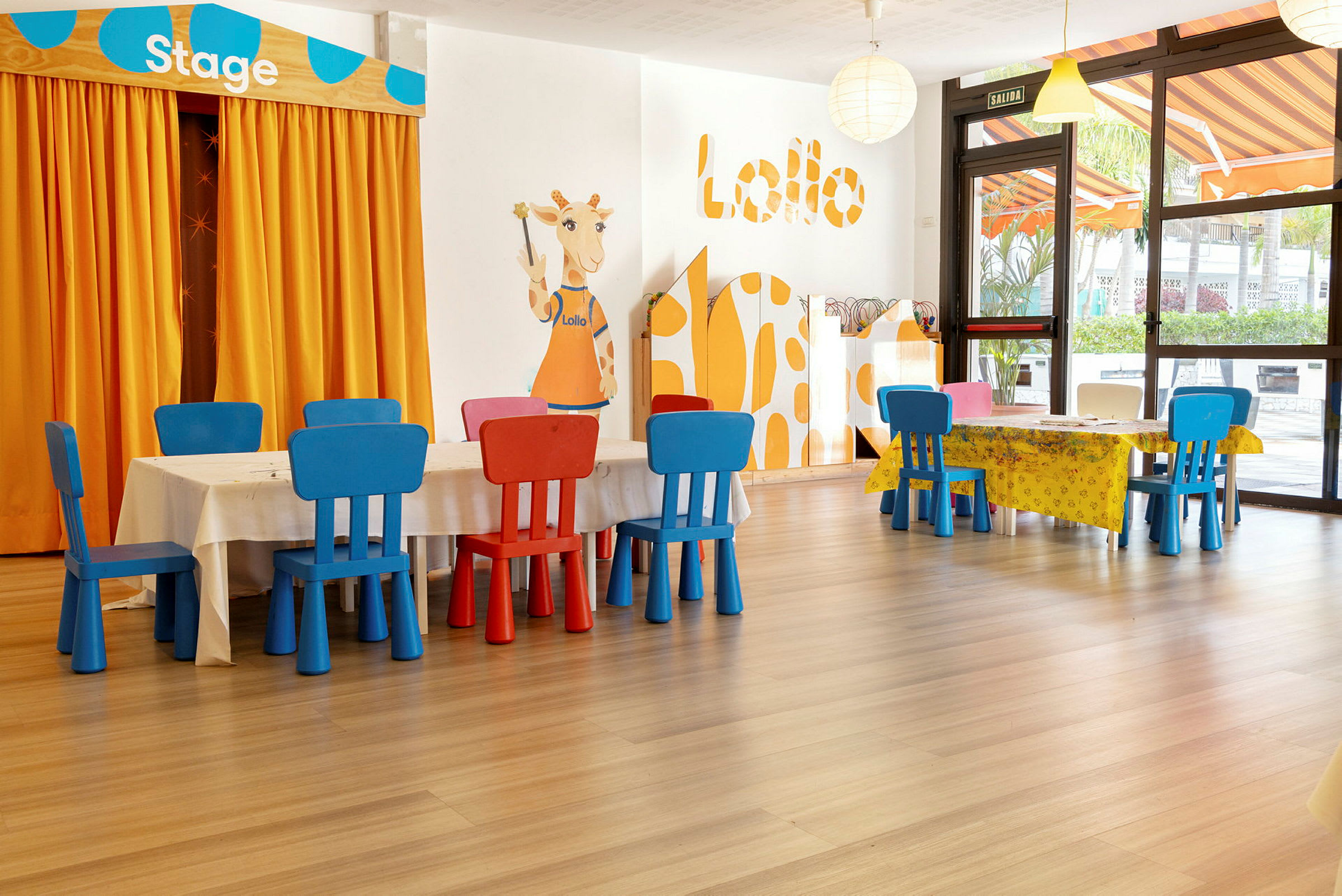 Ett barnvänligt rum med små bord och färgglada stolar. En illustration av en giraff vid en orange gardin märkt "Stage". (AI-genererad text)