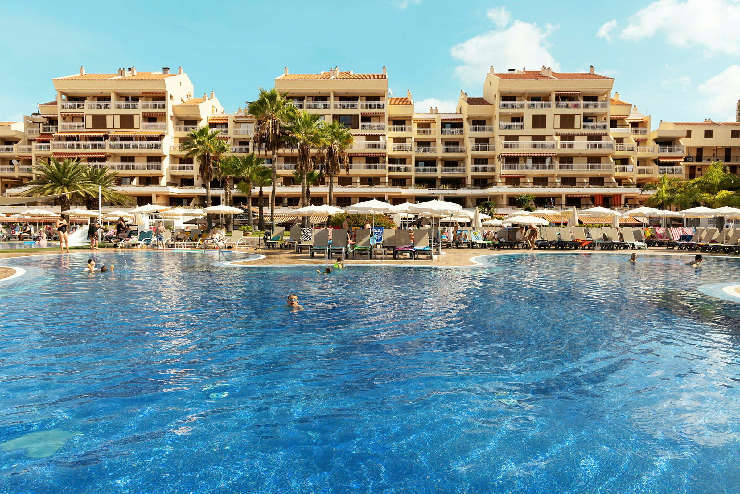 En stor pool omgiven av solstolar och parasoller framför en hotellbyggnad med flera våningar. (AI-genererad text)