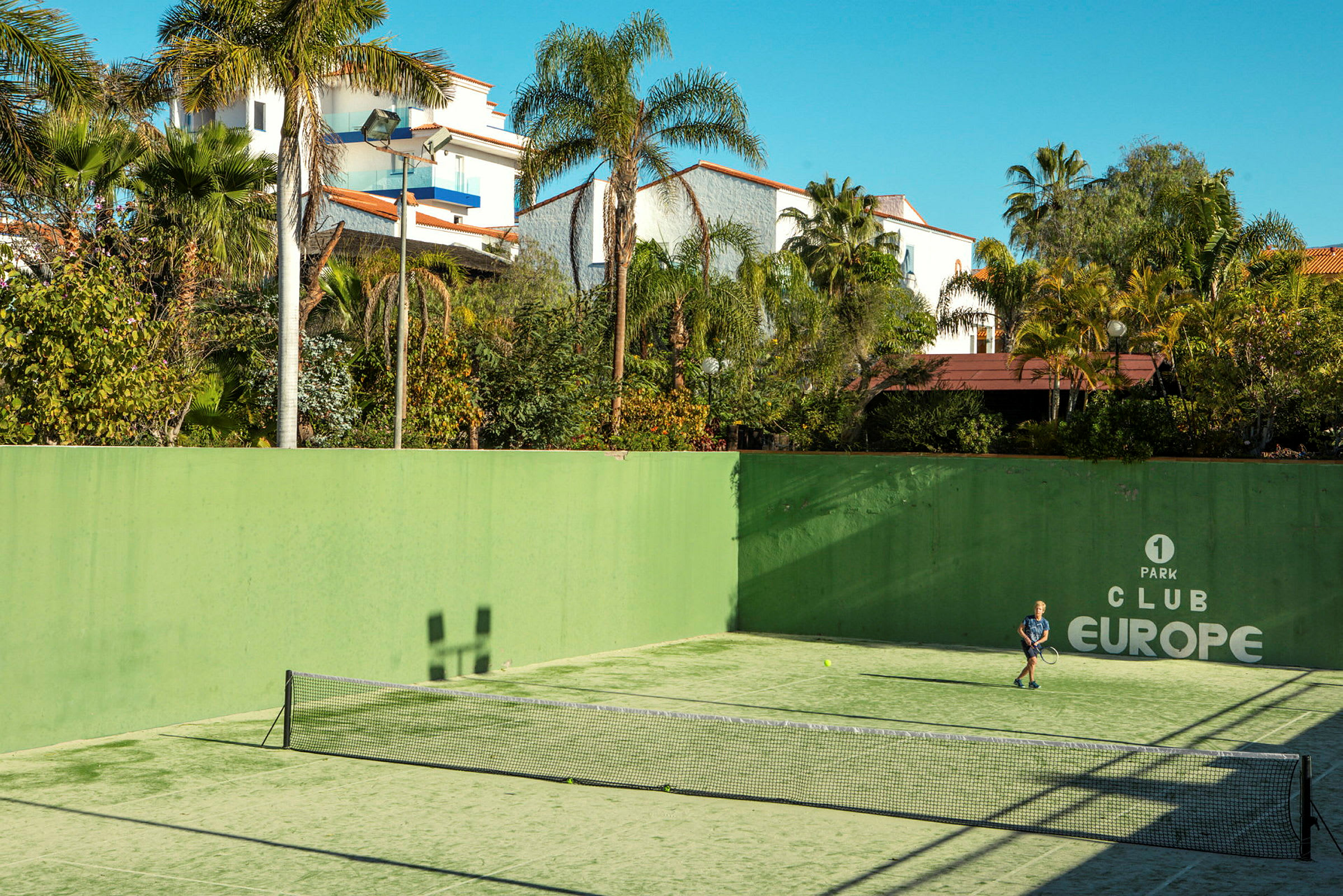En tennisbana utomhus med en person som spelar tennis, omgiven av gröna växter och byggnader. (AI-genererad text)