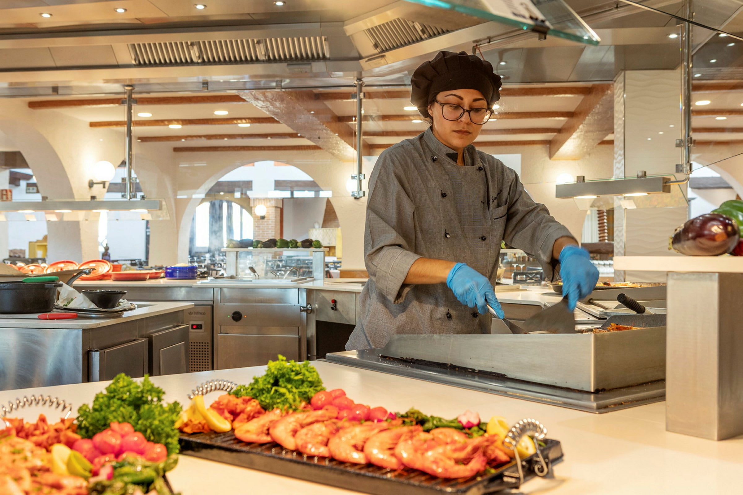 En kock tillagar mat vid en bufféstation i ett modernt kök med högar av färska ingredienser. (AI-genererad text)