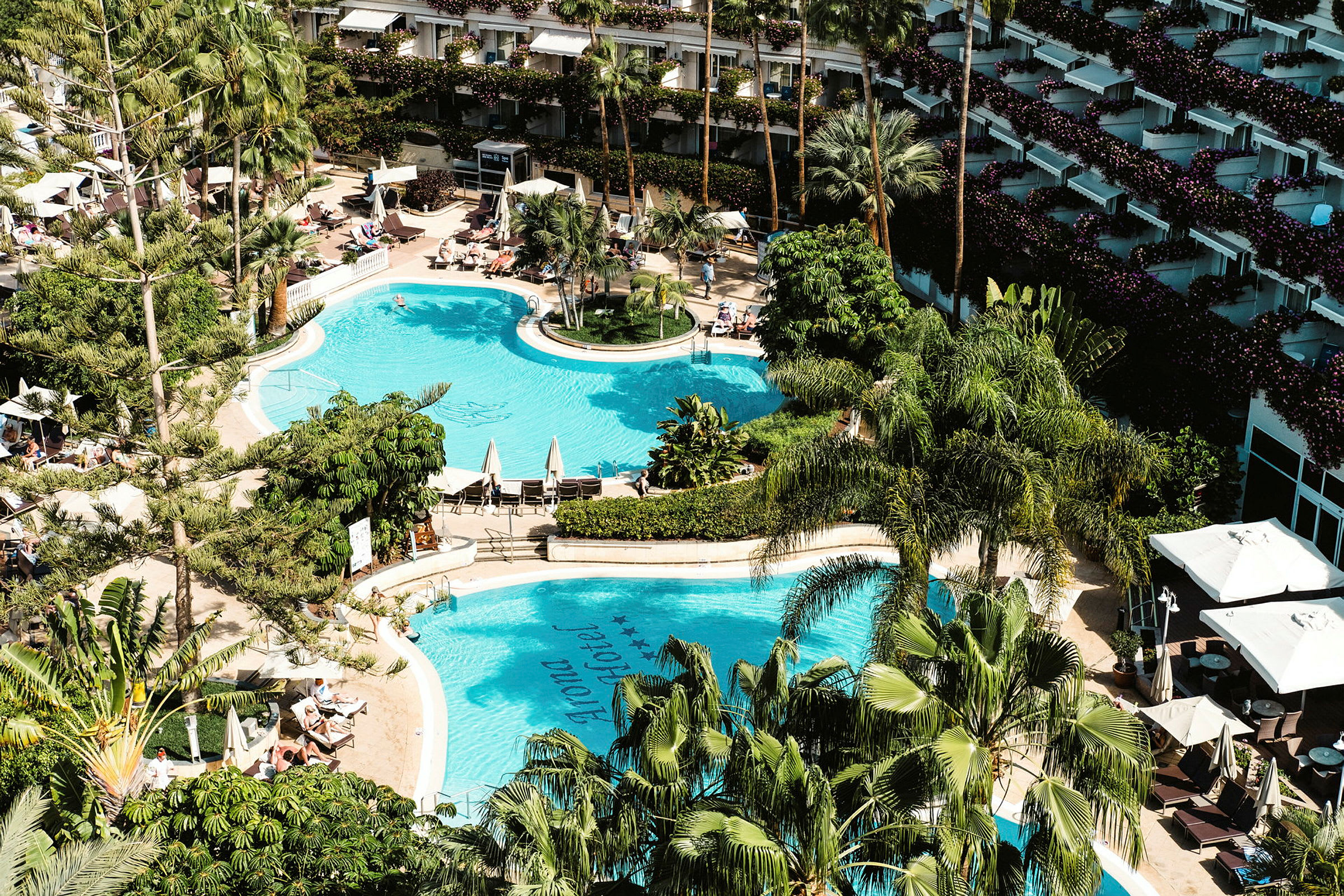 Flygbild av en hotellpoolområde omgiven av träd och solstolar, med en byggnad i bakgrunden. (AI-genererad text)