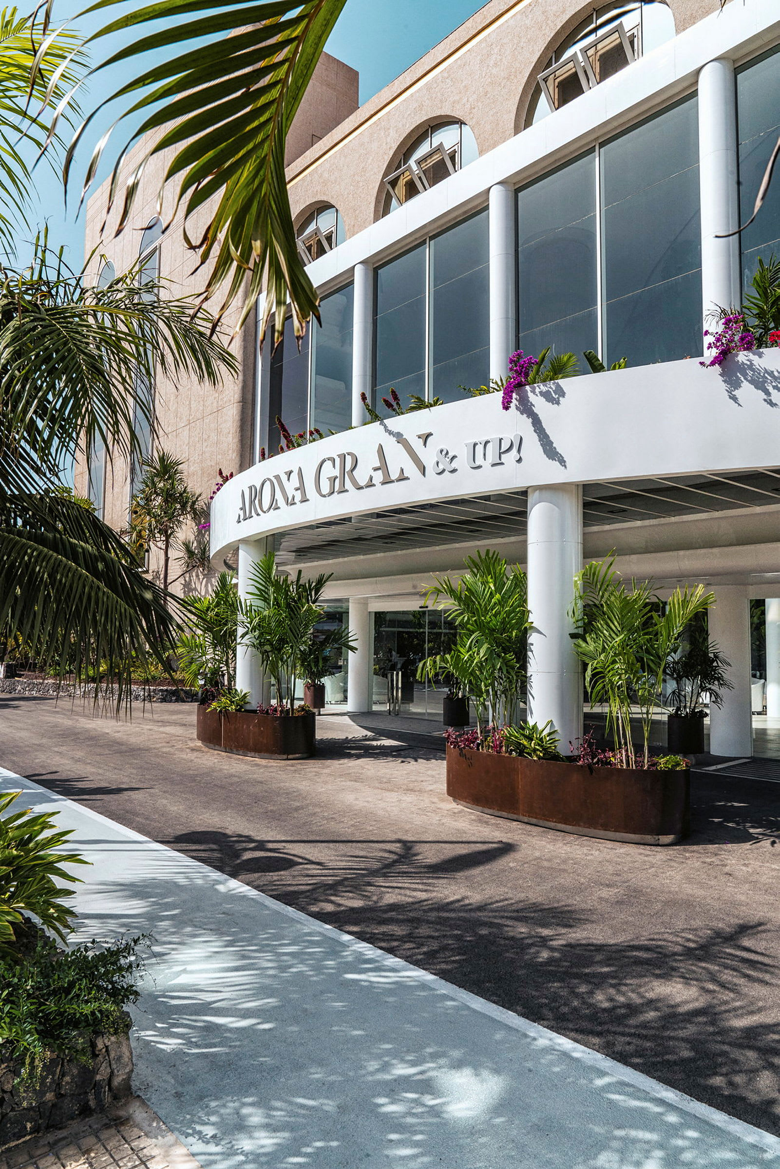 Hotellbyggnad med stora glasfönster, omgiven av växter och palmer. (AI-genererad text)