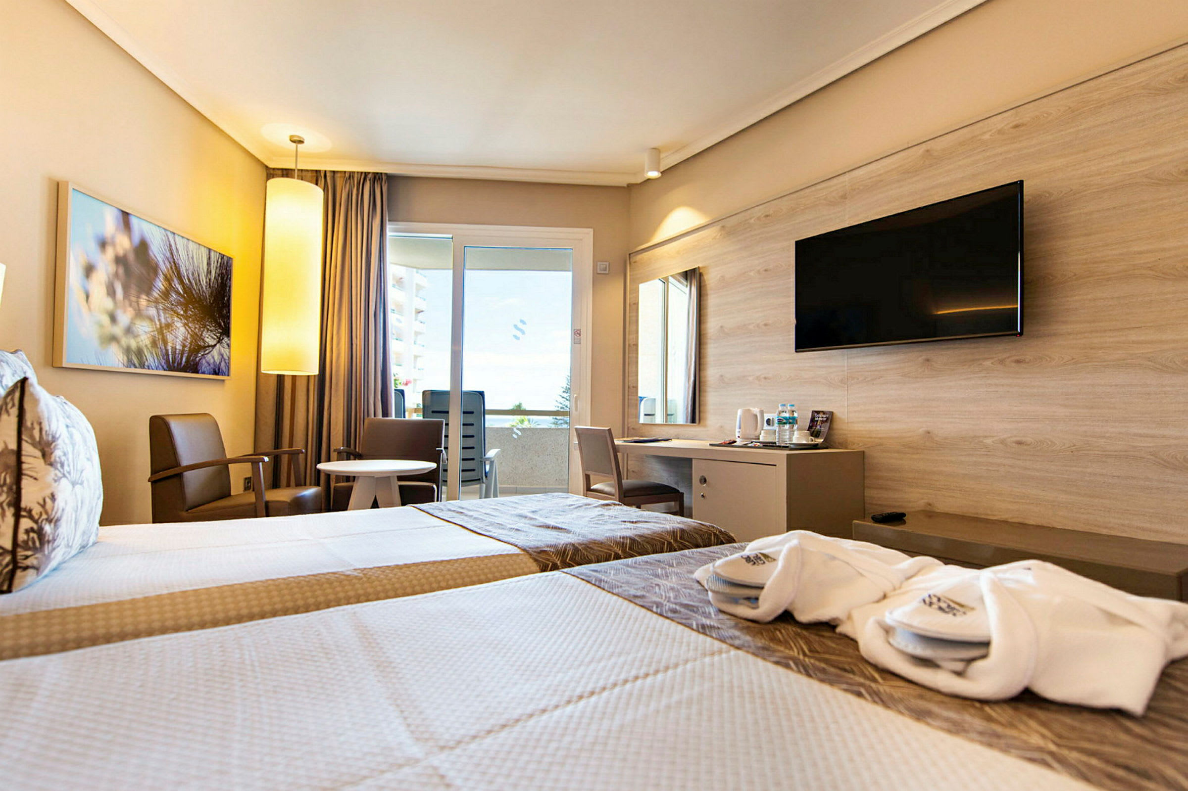 Ett hotellrum med två enkelsängar, sittmöbler vid ett runt bord, samt en skrivbordshörna med en väggmonterad TV ovanför. (AI-genererad text)