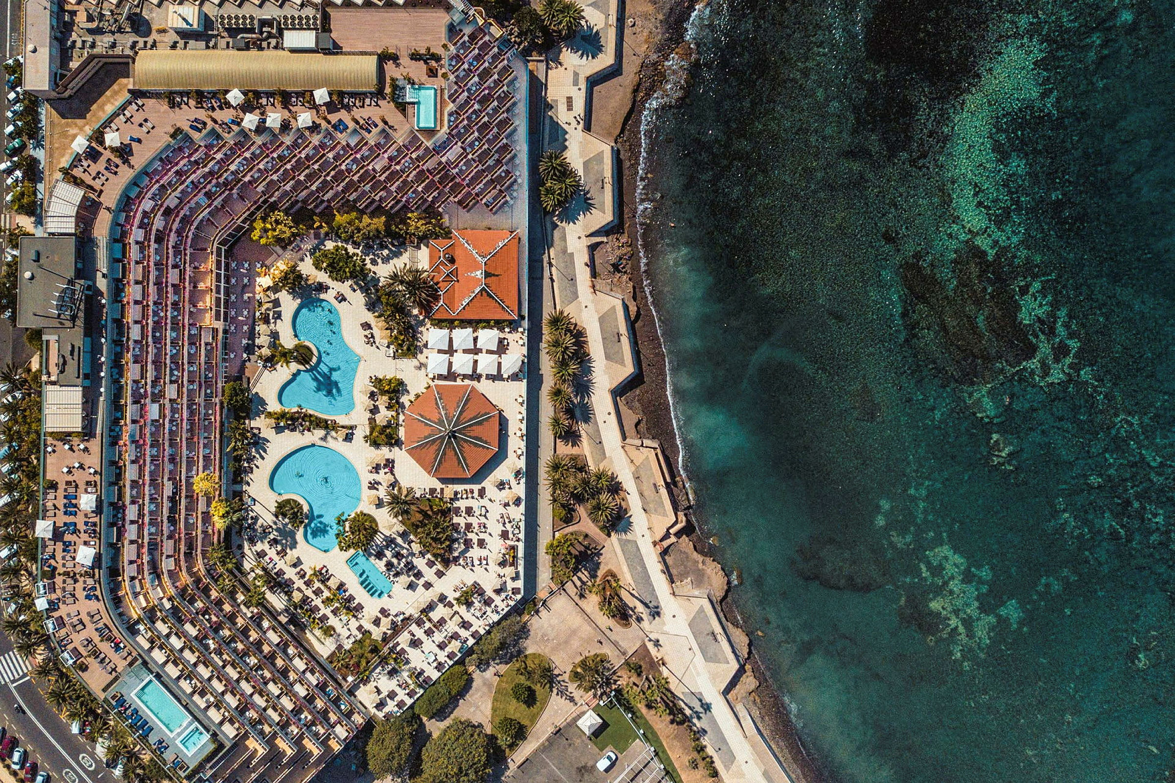 Flygbild av hotellområde med pooler, solstolar och hav vid sidan. (AI-genererad text)