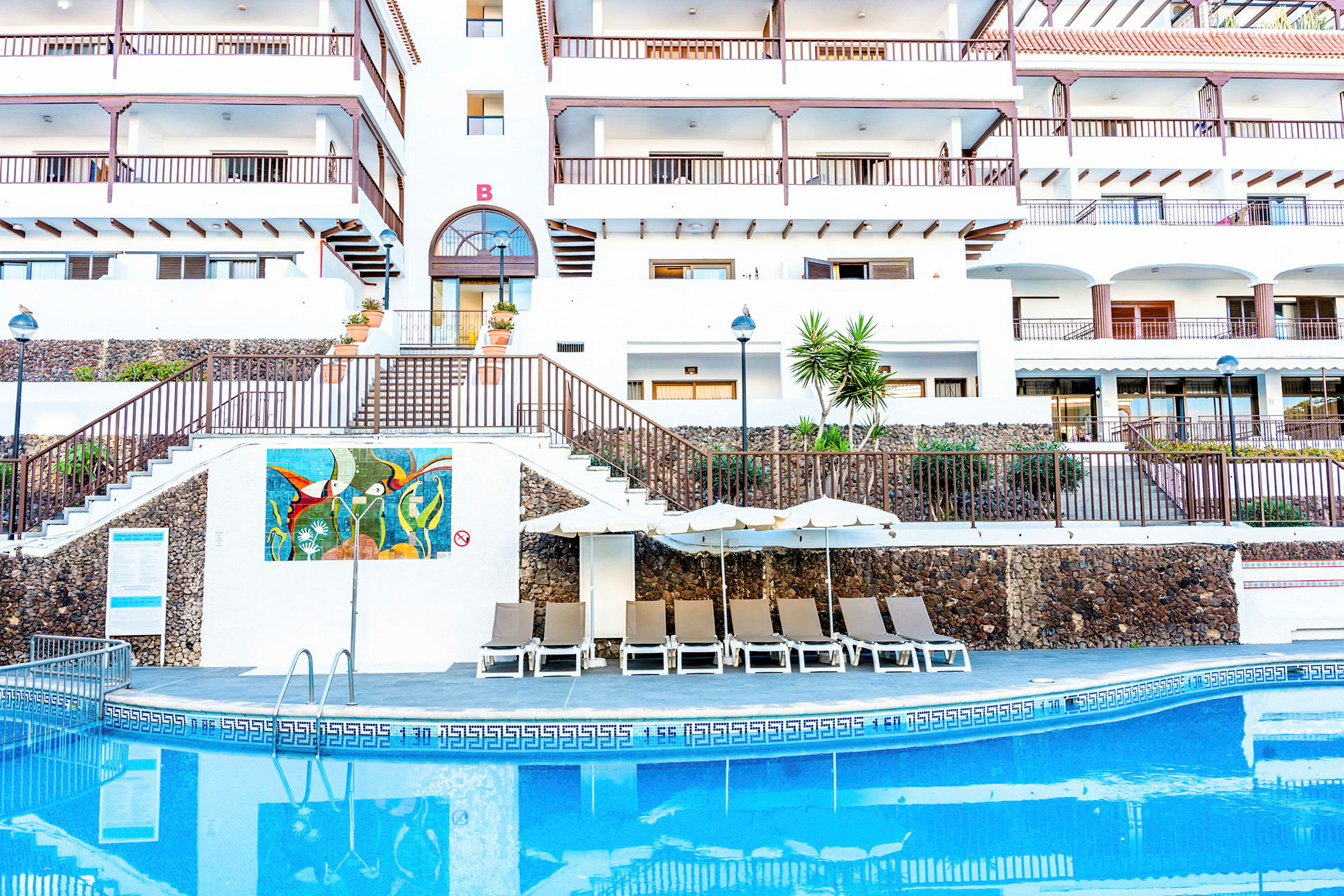 En byggnad med en pool och solstolar framför. (AI-genererad text)
