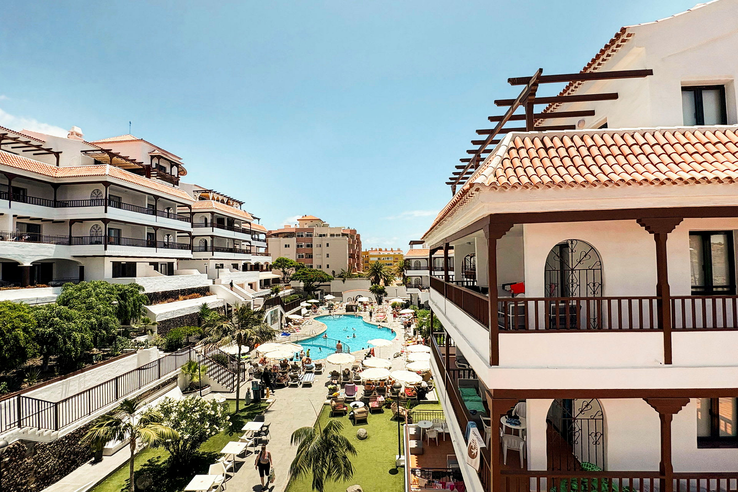 Bild av en hotellanläggning med flera våningar och röd taktegel. I mitten syns en poolområde med solstolar och parasoller omgiven av grönska. (AI-genererad text)