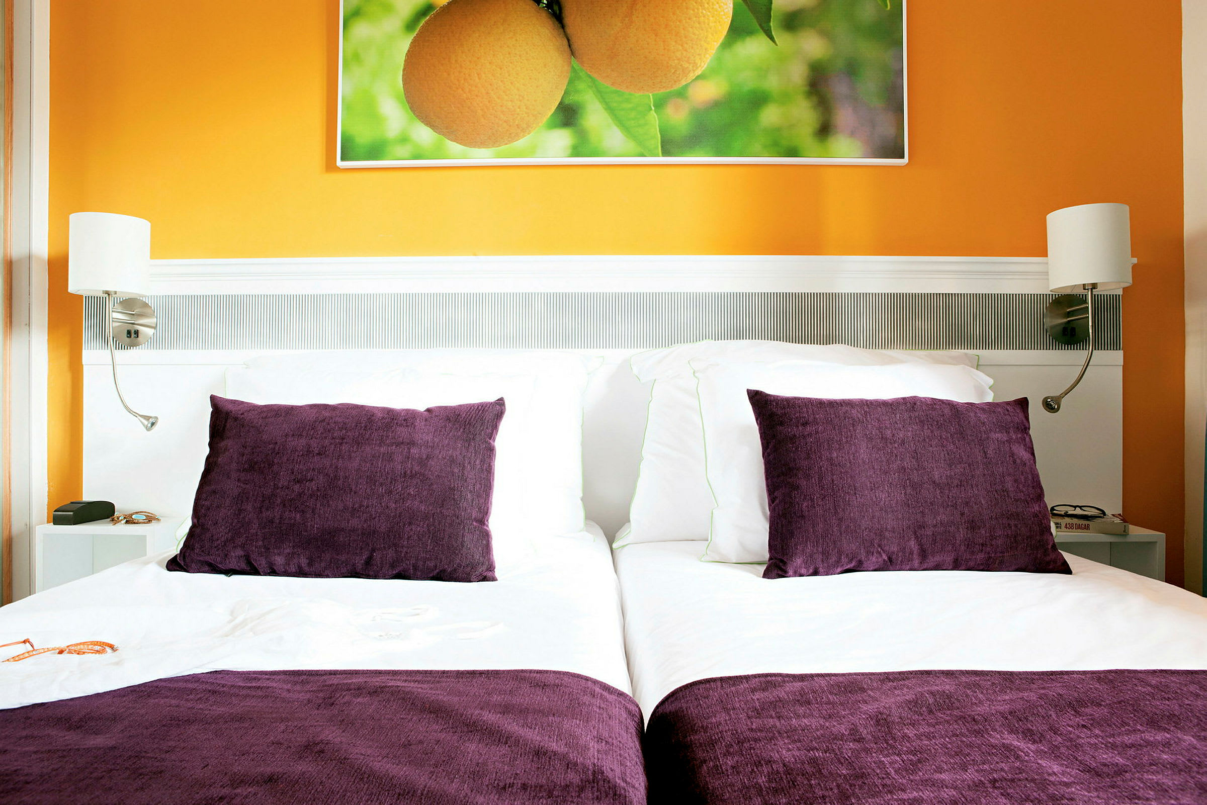 En säng med vita lakan och lila kuddar framför en orange vägg, där ett foto av apelsiner hänger. (AI-genererad text)