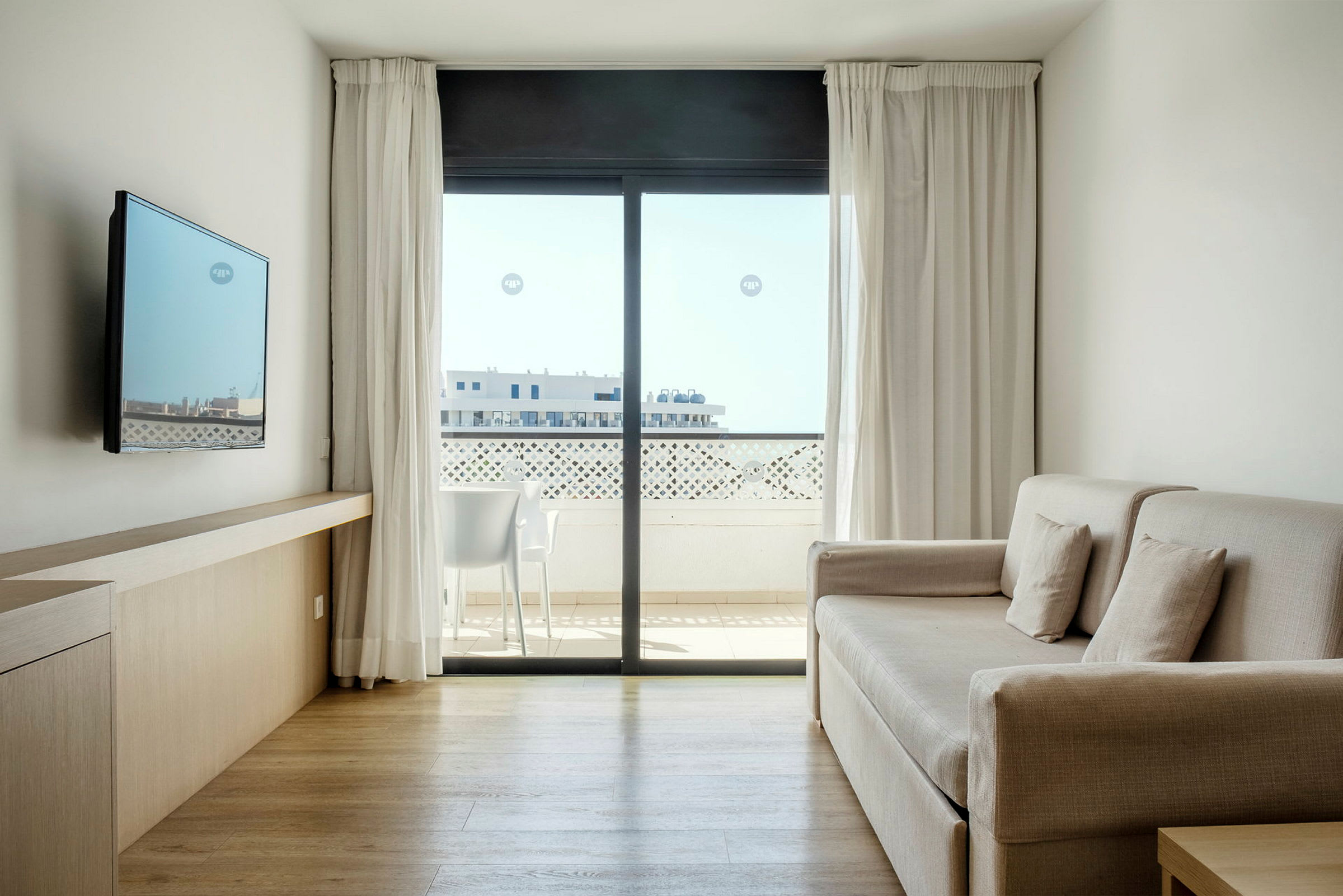 En ljus och modern hotellrum med soffa, väggmonterad TV och stort fönster med utsikt mot en balkong. (AI-genererad text)