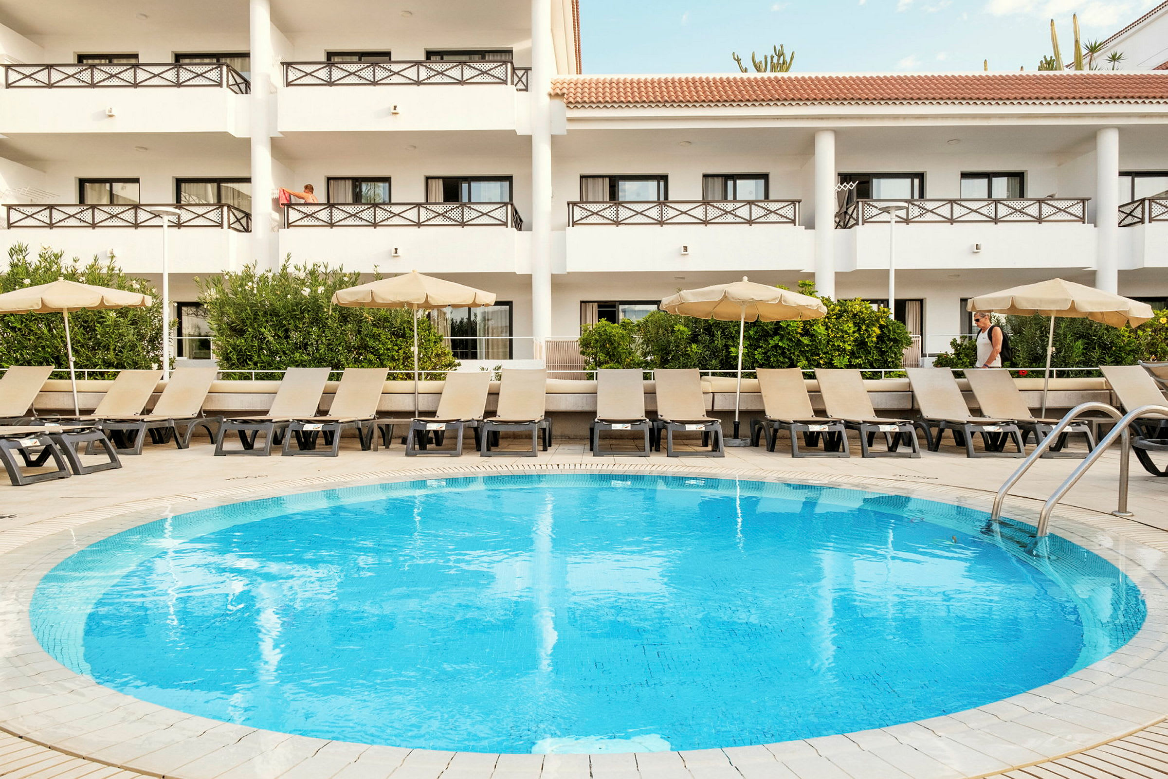Bild av en rund pool med solstolar och parasoller. Bakom poolområdet syns en byggnad med balkonger. (AI-genererad text)