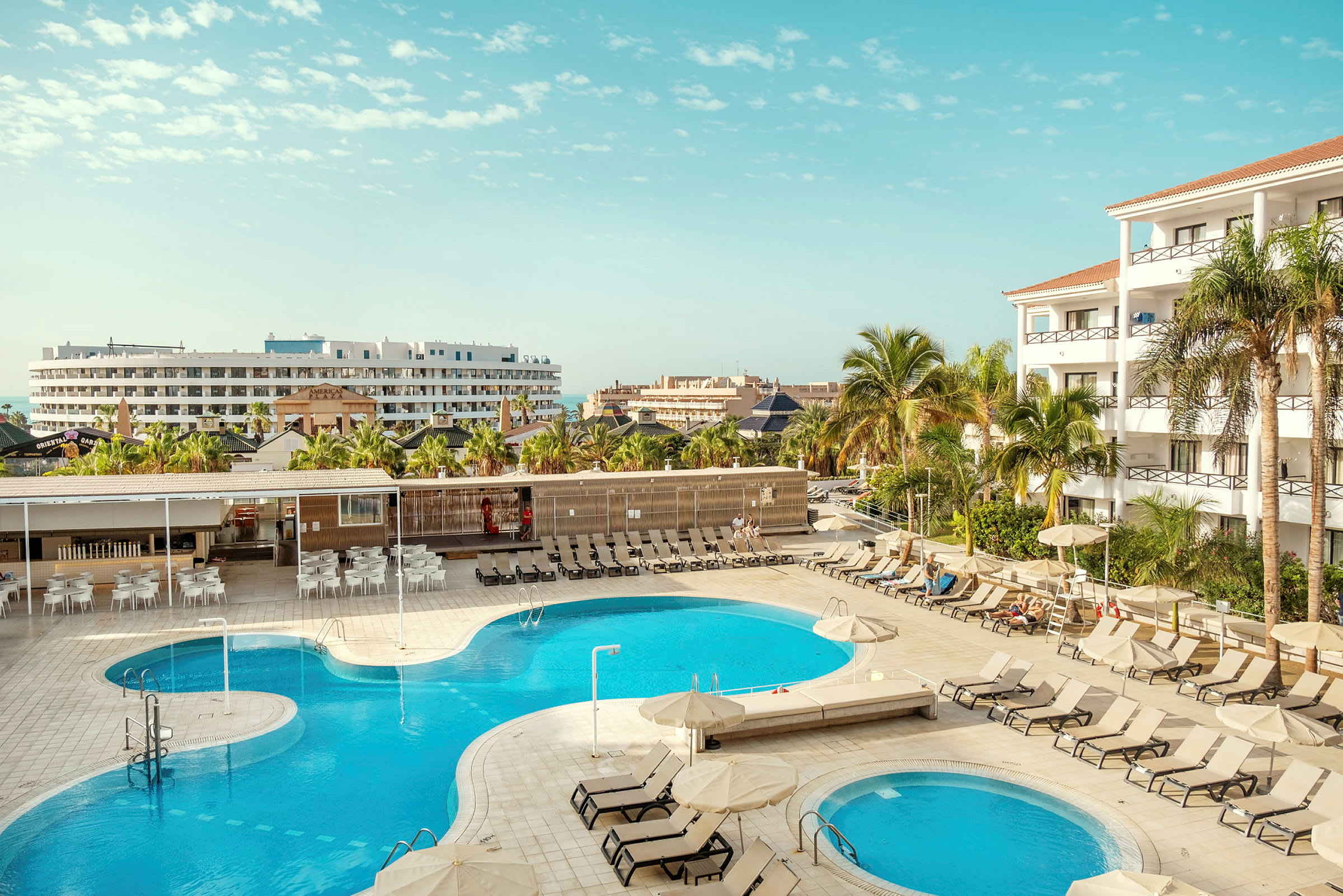 En hotellpool med omgivande solstolar och byggnader i bakgrunden. (AI-genererad text)