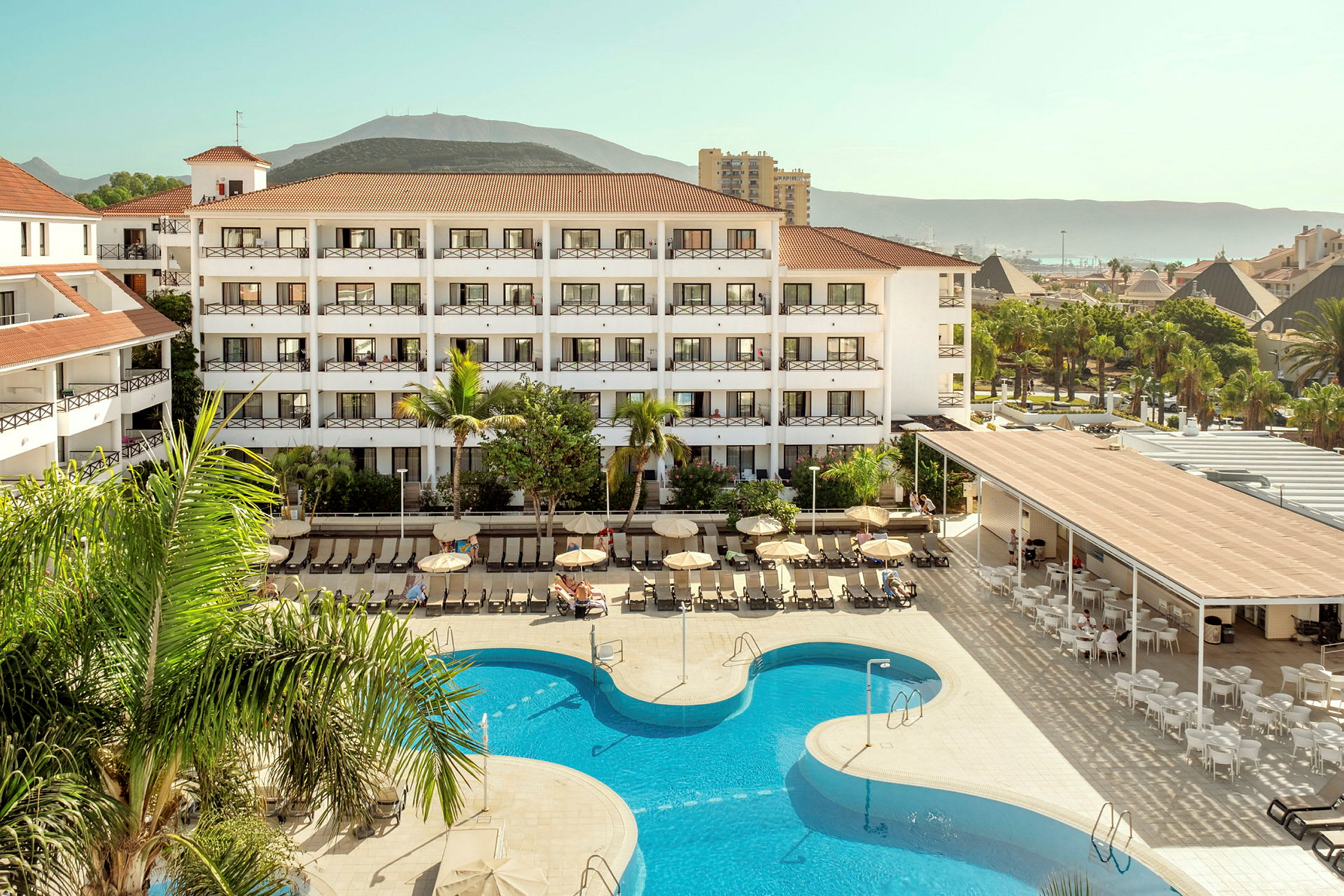Hotellkomplex med bassäng och solstolar, omgiven av grönska och berg i bakgrunden. (AI-genererad text)