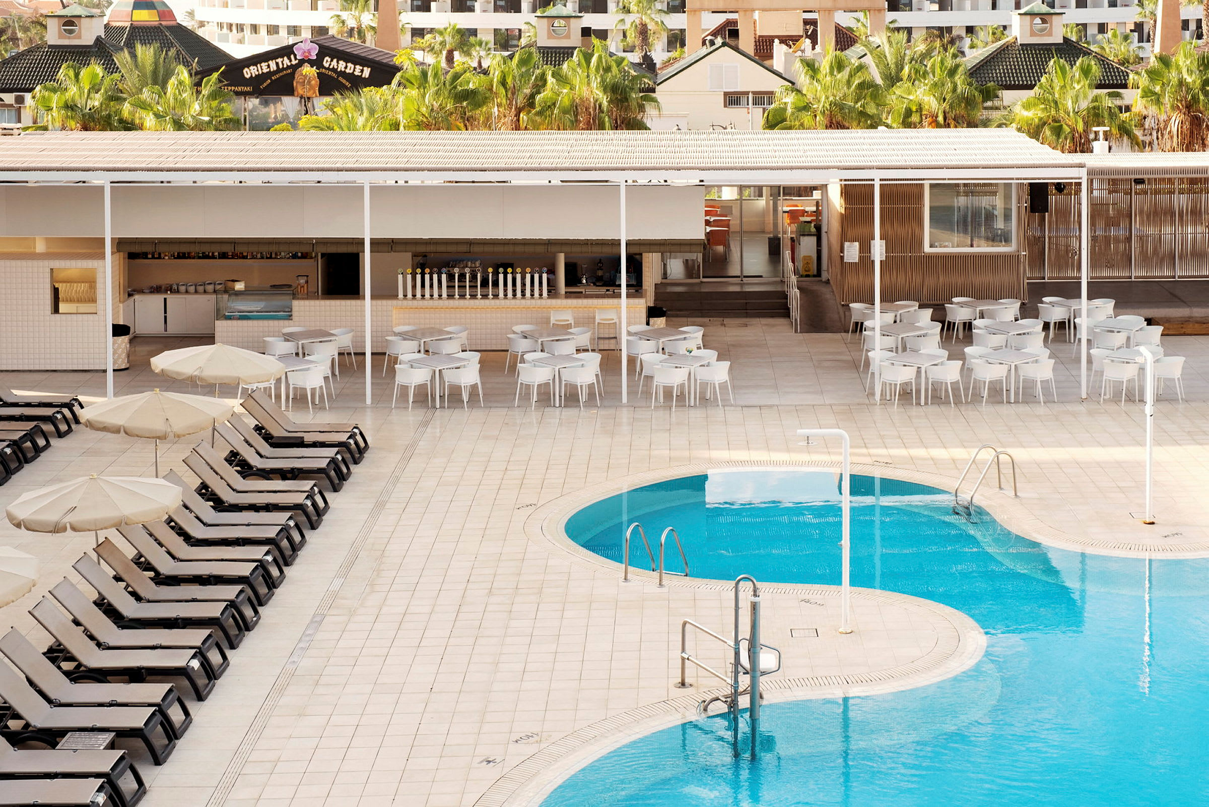 Bild av en pool med solsängar och parasoller i förgrunden, samt en bar och sittplatser i bakgrunden. (AI-genererad text)