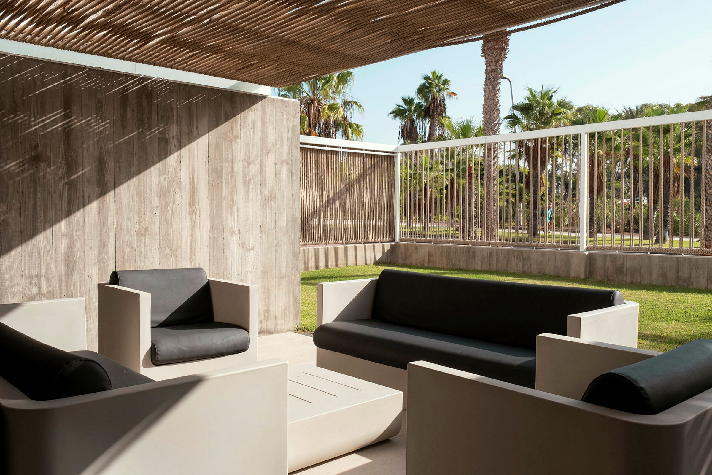 Utemöbler med svart sittdyna under en träpergola, med utsikt över grönt gräs och palmer. (AI-genererad text)