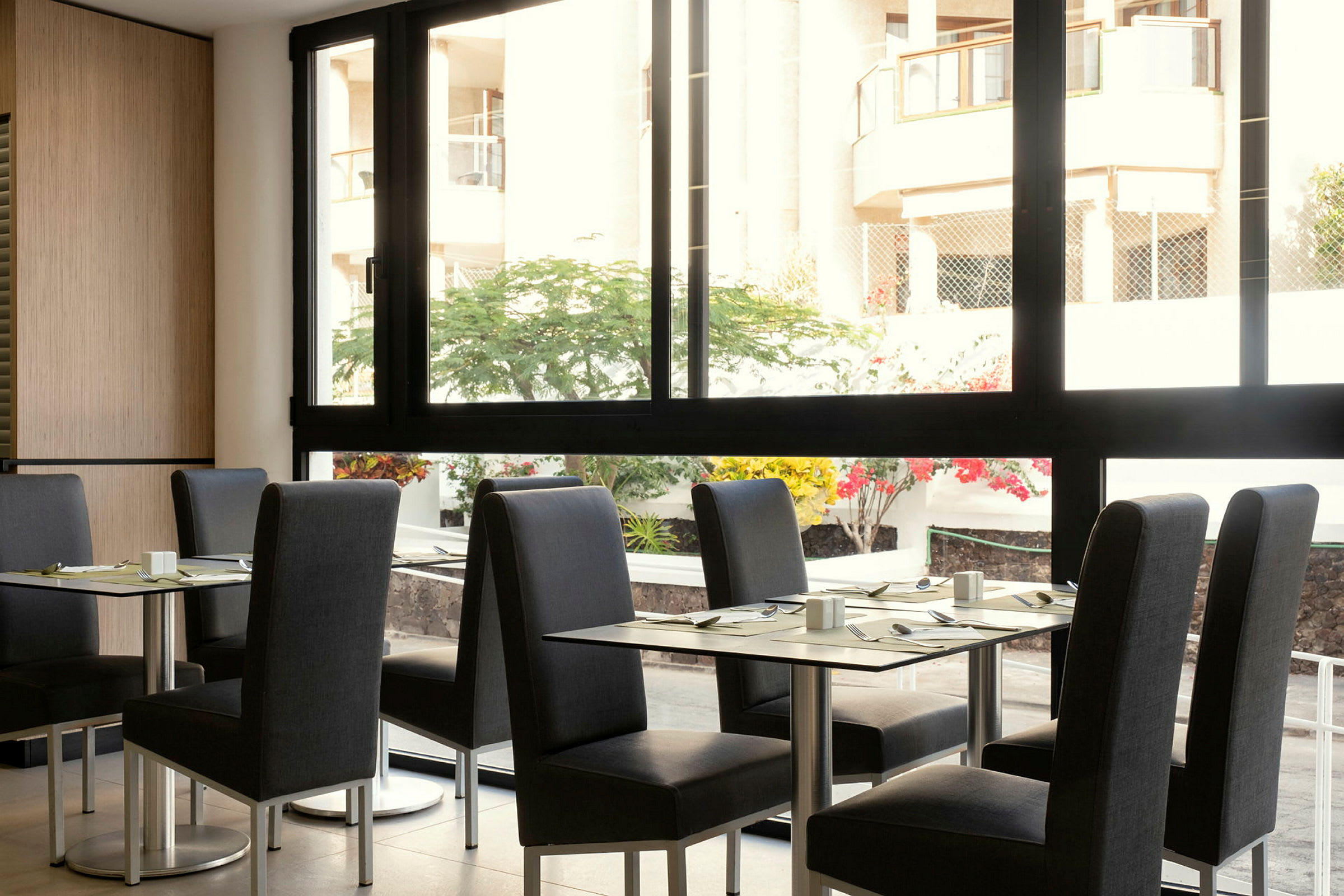 En restaurang med bord och stolar, med utsikt mot en gårdsplan genom en stor fönsterruta. (AI-genererad text)