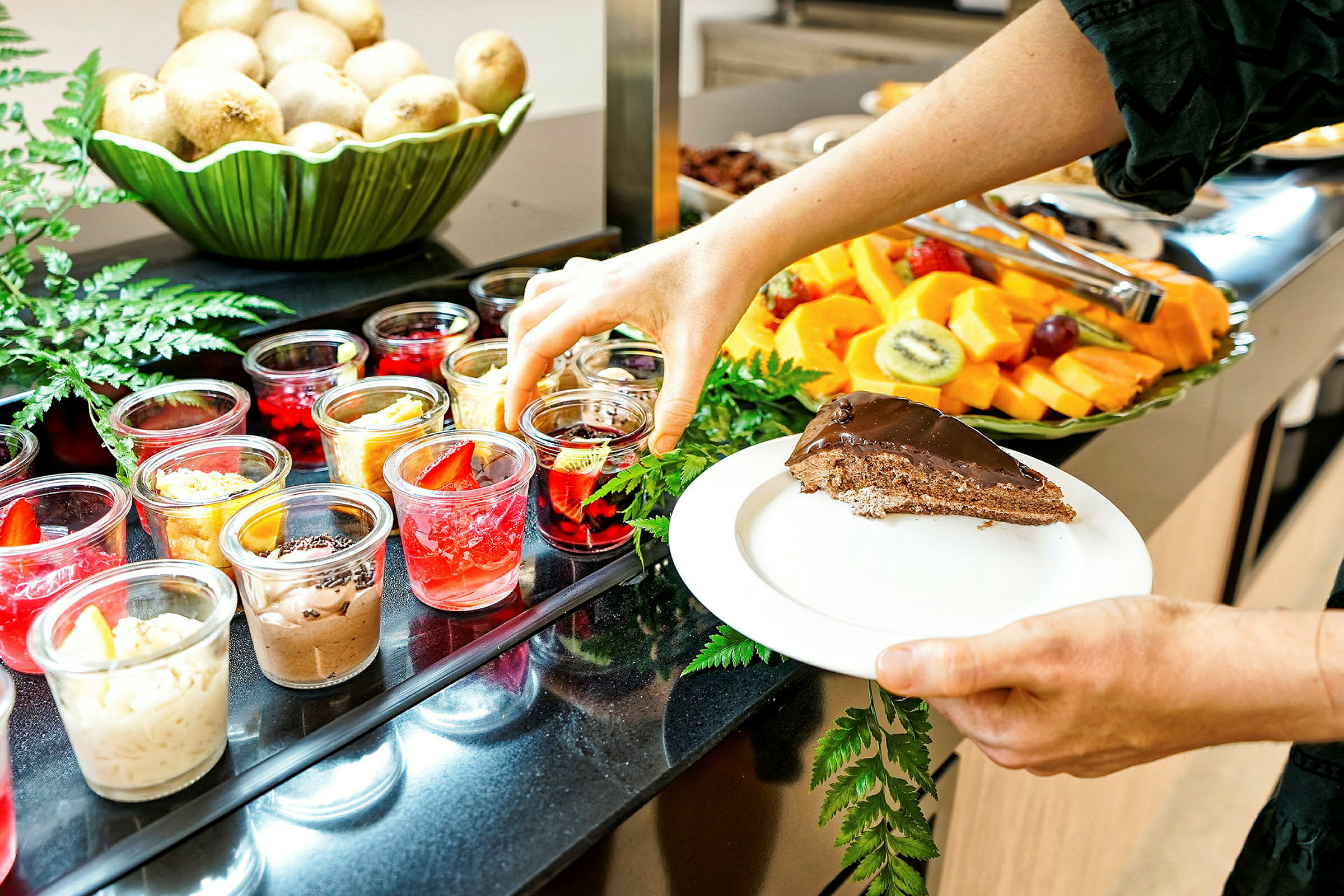 En person tar en efterrätt från en buffé med ett urval av små desserter i glas, frukter och en skiva chokladkaka på en tallrik. (AI-genererad text)