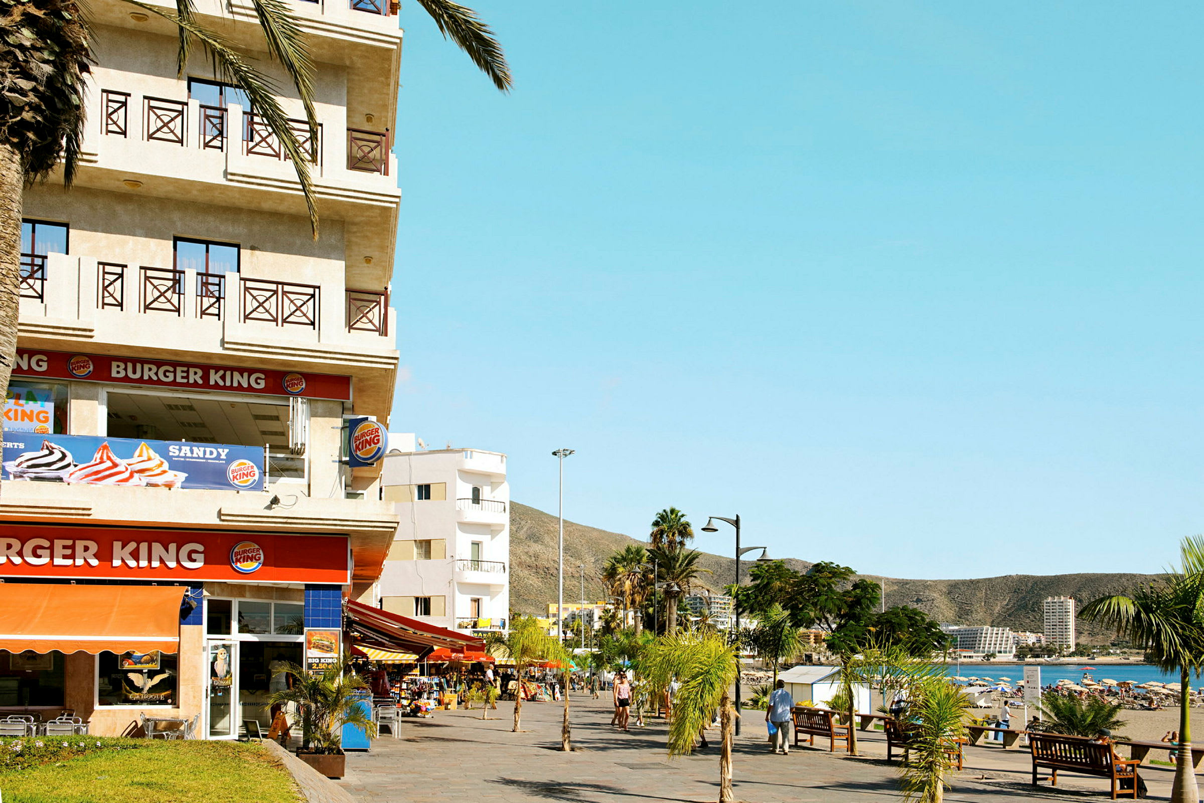 Bilden visar en Burger King-restaurang belägen nära en strandpromenad, med palmer och människor som promenerar. (AI-genererad text)