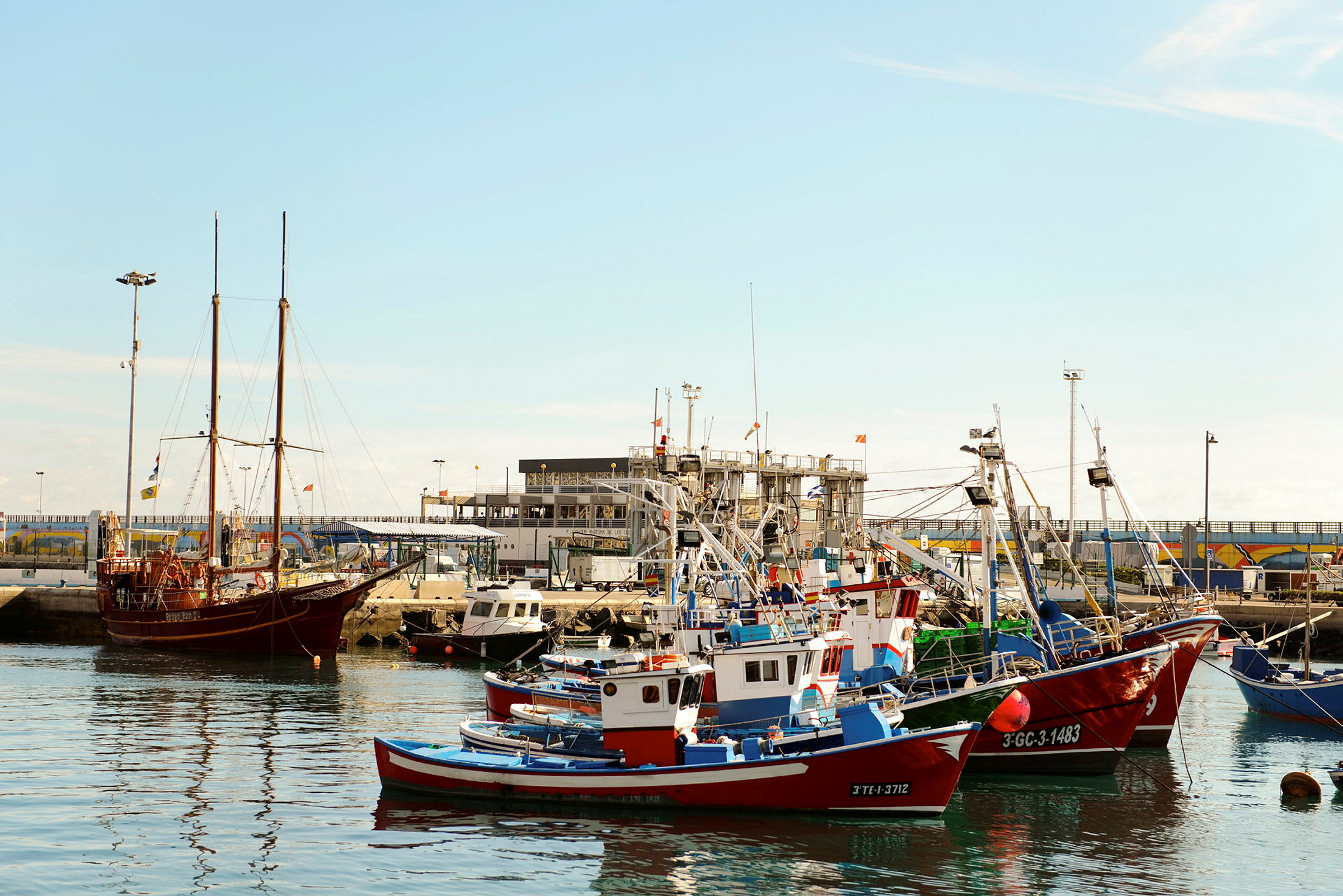 Bilden visar en hamn med flera fiskebåtar och en större segelbåt vid kajen. (AI-genererad text)