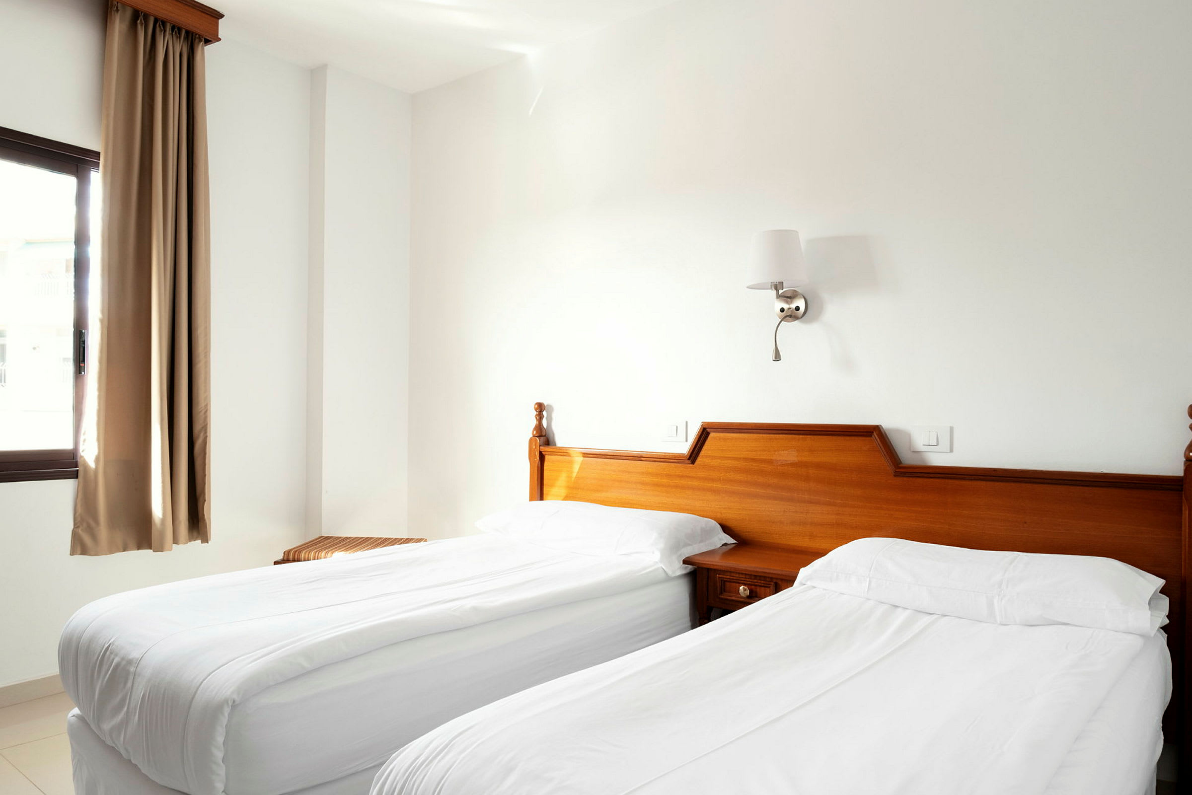 Ett hotellrum med två enkelsängar klädda i vita sängkläder, trämöbler och en fönster med beige gardiner. (AI-genererad text)