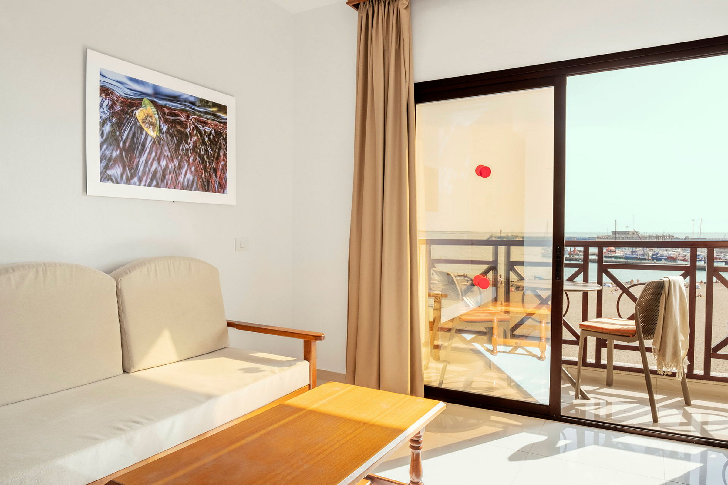Utsikt från ett hotellrum med en beige soffa, ett träbord och en balkong med utsikt över stranden. (AI-genererad text)