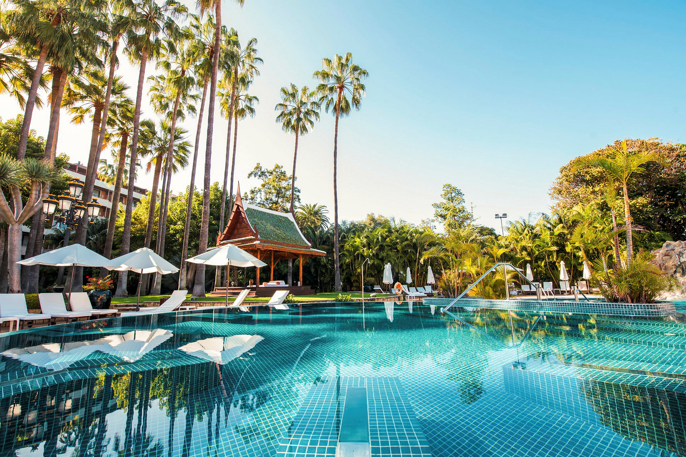 En swimmingpool omgiven av solstolar, parasoller och palmer med ett thailändskt paviljong i bakgrunden. (AI-genererad text)