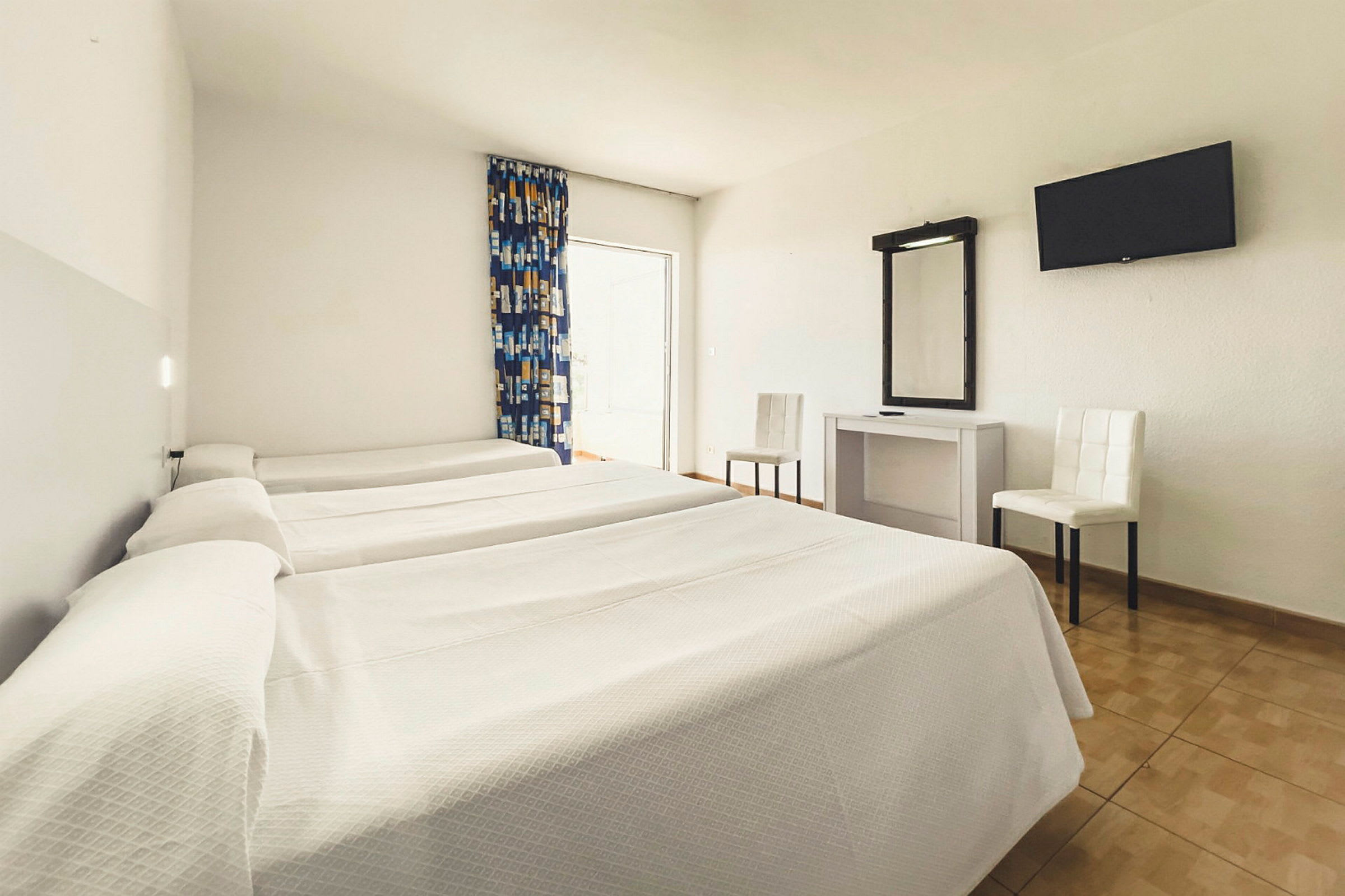Ett hotellrum med tre enkelsängar, ett skrivbord med spegel, två stolar och en väggmonterad TV. (AI-genererad text)