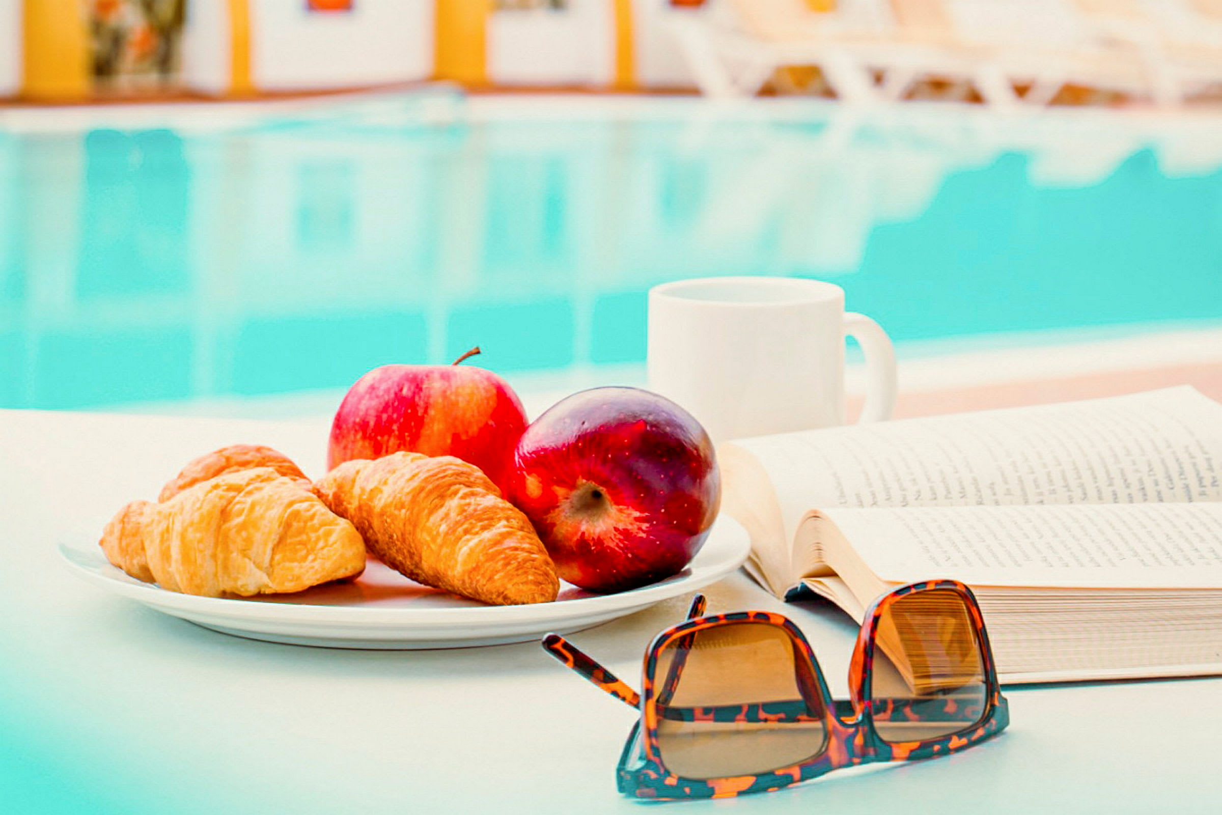 En tallrik med croissanter och äpplen står på ett bord bredvid en bok och ett par solglasögon. I bakgrunden syns en pool. (AI-genererad text)