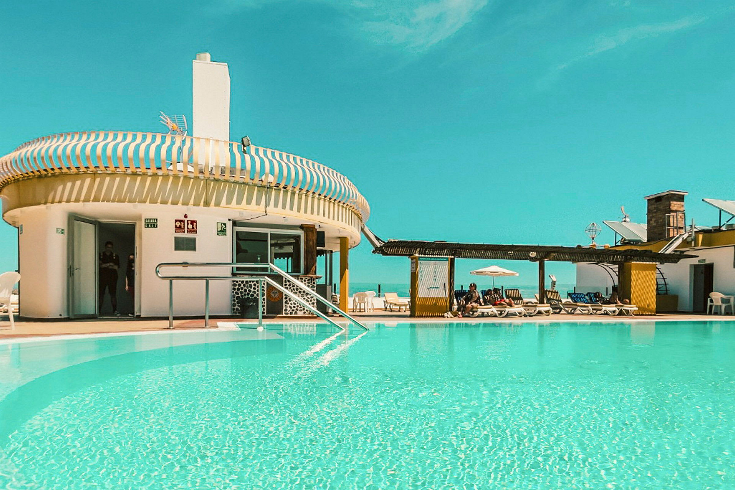 En utomhuspool vid en byggnad med solstolar och ett klart blått himmel. (AI-genererad text)