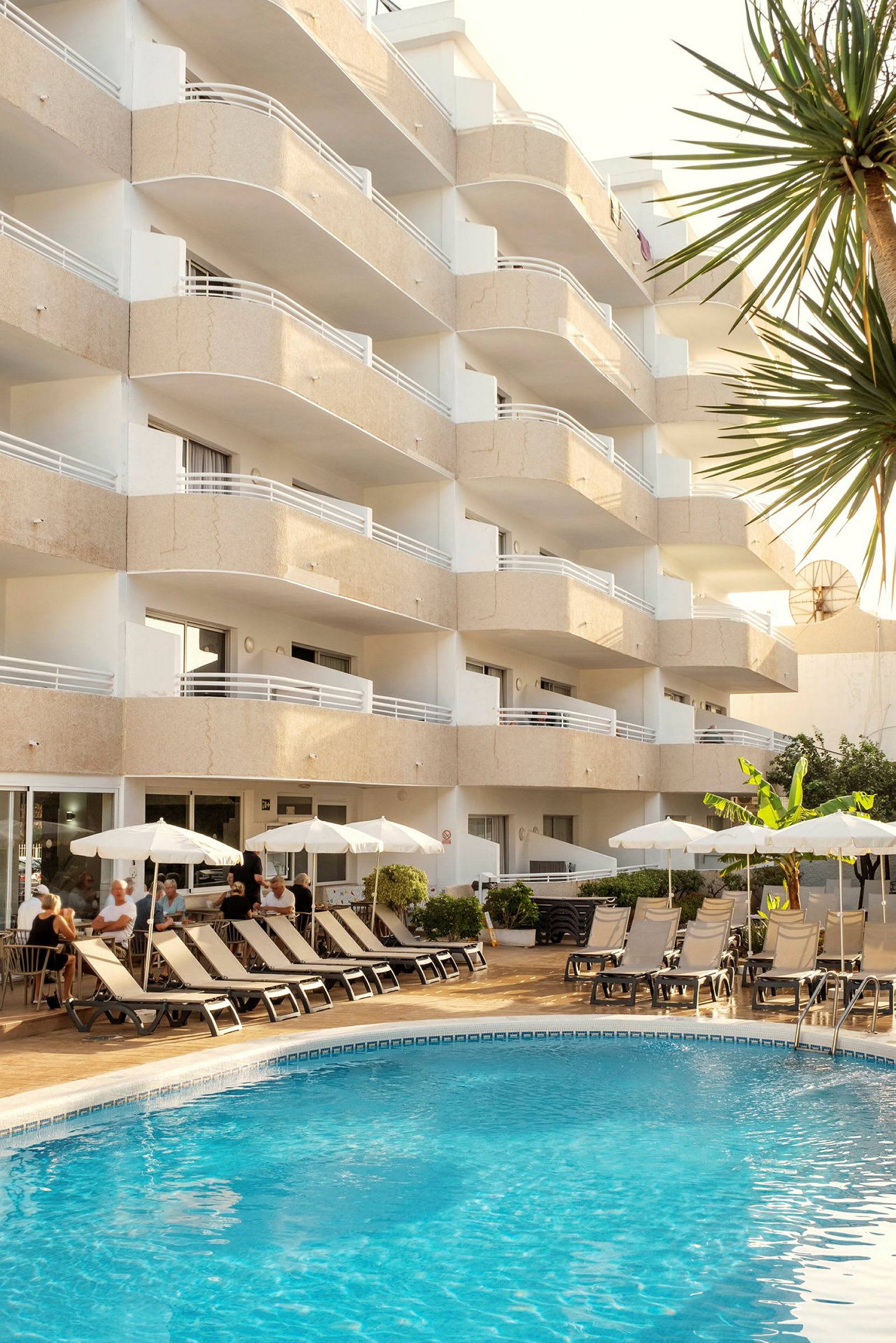 Hotellfasad med balkonger och en utomhuspool framför. Solstolar och parasoll står på poolområdet som omges av grönska. (AI-genererad text)