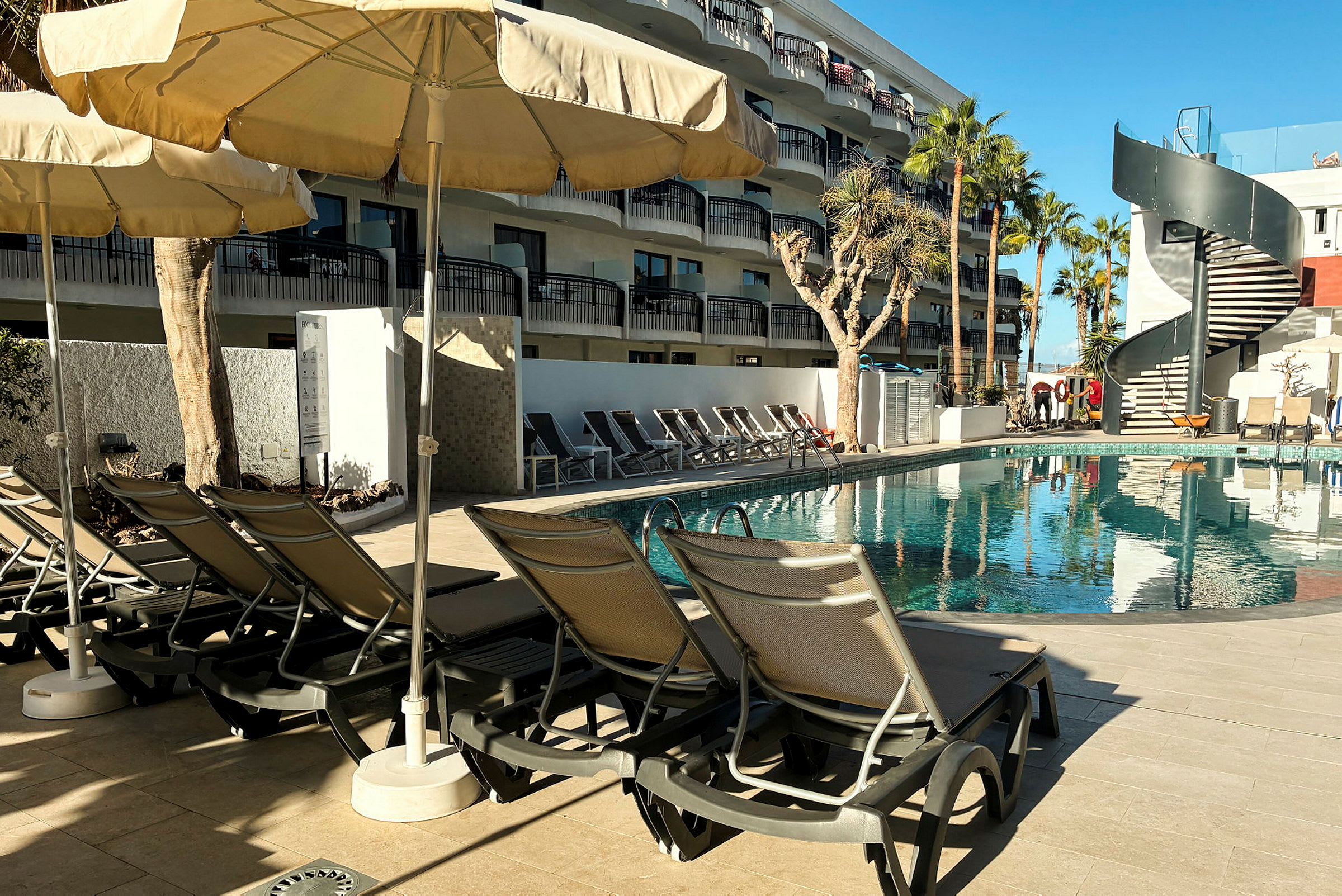 Bild av en pool med solstolar och parasoller bredvid samt en byggnad med balkonger i bakgrunden. (AI-genererad text)