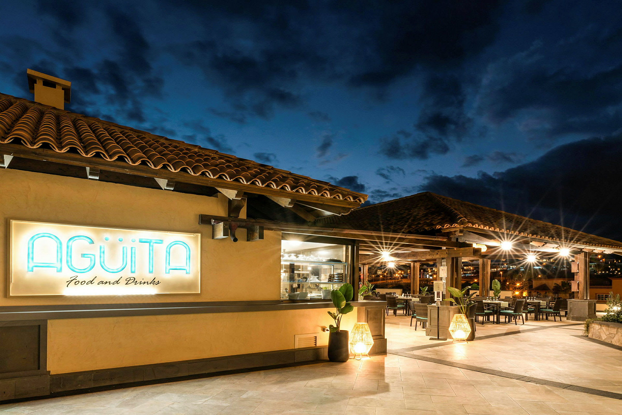 Exteriörbild av en restaurang med terrakottatak och namnet Agüita upplyst på en skylt. (AI-genererad text)