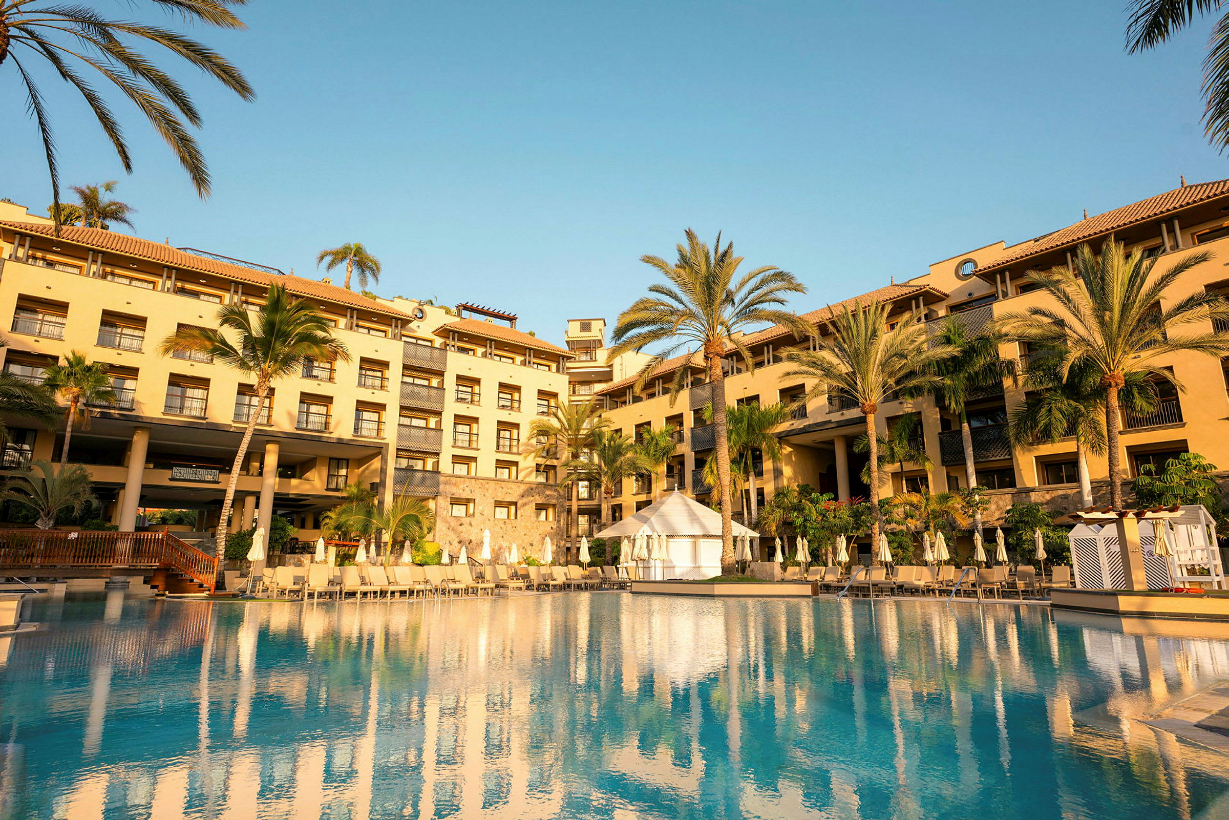 Hotellbyggnad med pool framför byggnaden, omgiven av palmer och klar blå himmel. (AI-genererad text)