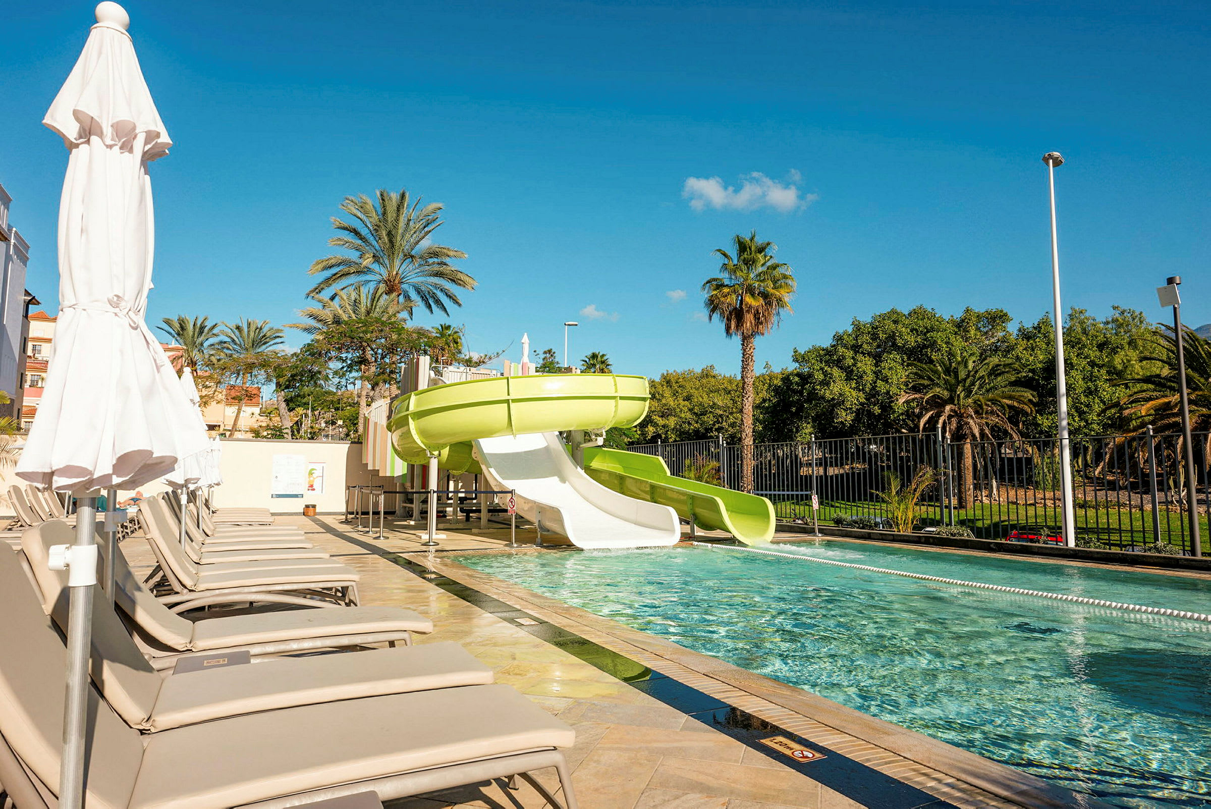 Gröna vattenrutschbanor vid en swimmingpool omgiven av solstolar och palmer, med klar blå himmel. (AI-genererad text)