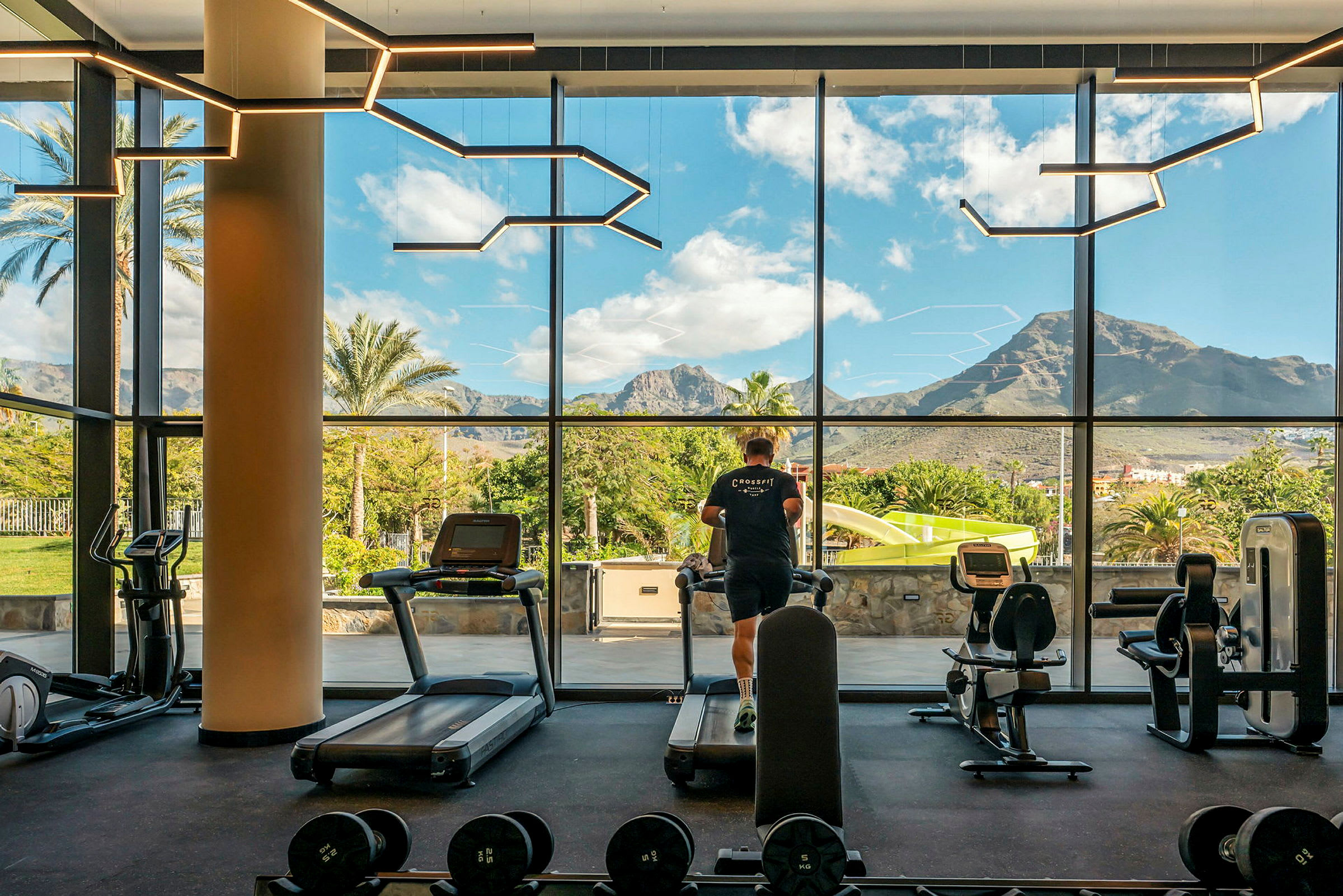 Utsikt från ett gym med stora fönster, som visar en person som springer på ett löpband. Utanför finns grönska, palmer och berg. (AI-genererad text)
