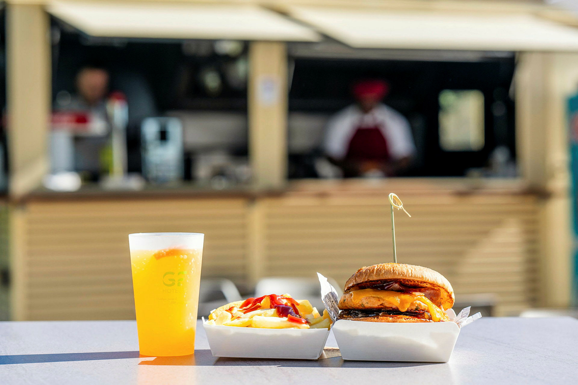Mat som består av en hamburgare, pommes frites och en dryck i en plastmugg på ett bord utomhus, med bakgrund av en matservering. (AI-genererad text)