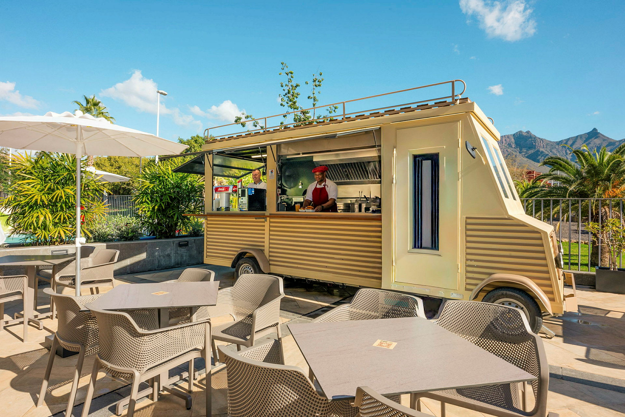 En gul foodtruck med serveringslucka är parkerad i en utomhusmiljö med bord, stolar och parasoll. (AI-genererad text)