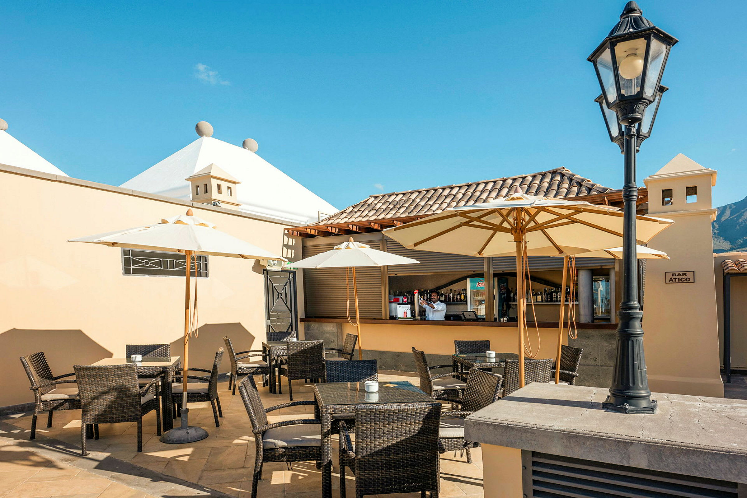 En utomhusrestaurang med bord, stolar och parasoller under en klarblå himmel. (AI-genererad text)