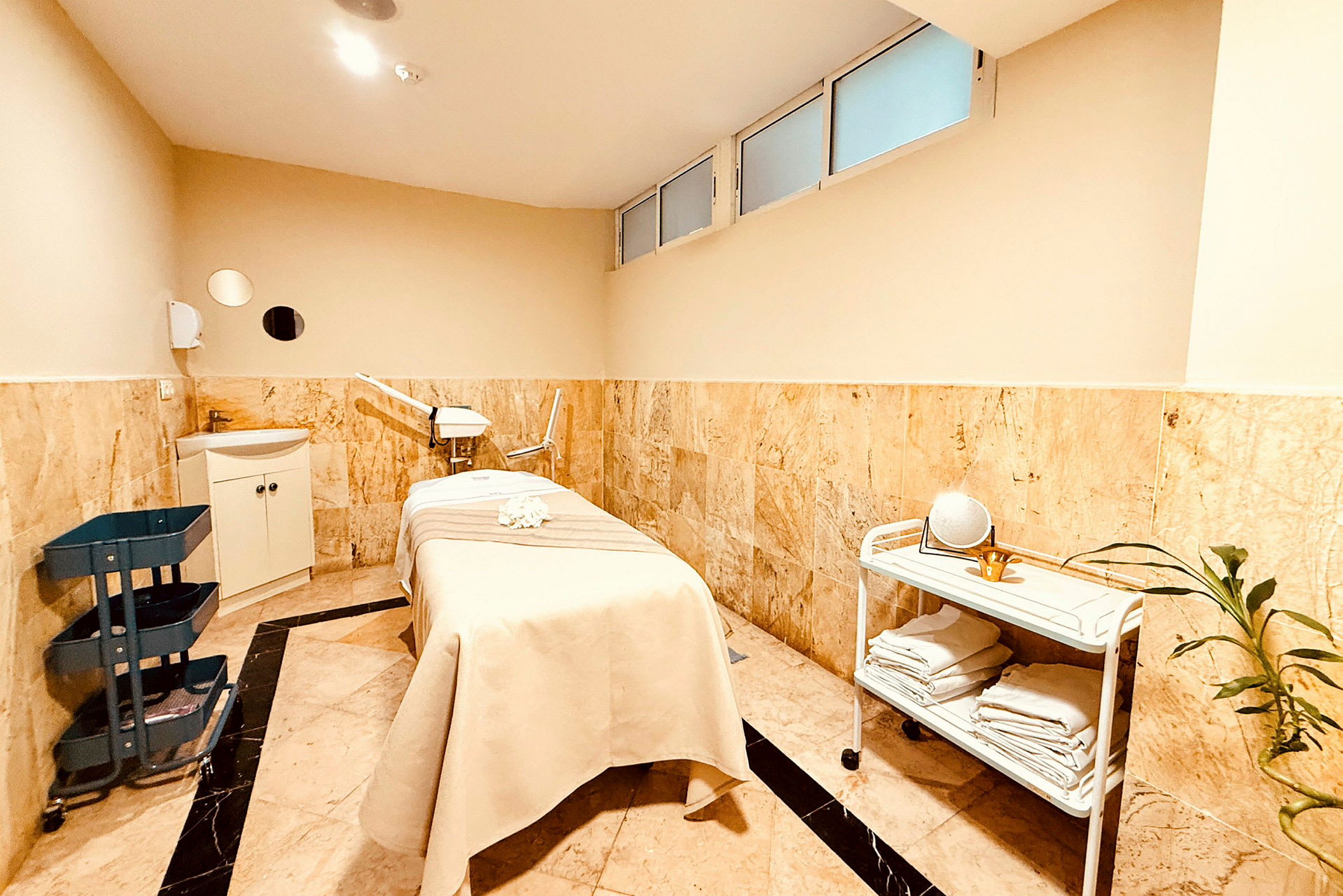 Ett rum med ett massagebord täckt av ett beige lakan, några hyllor som förvarar handdukar och en liten planta i hörnet. (AI-genererad text)
