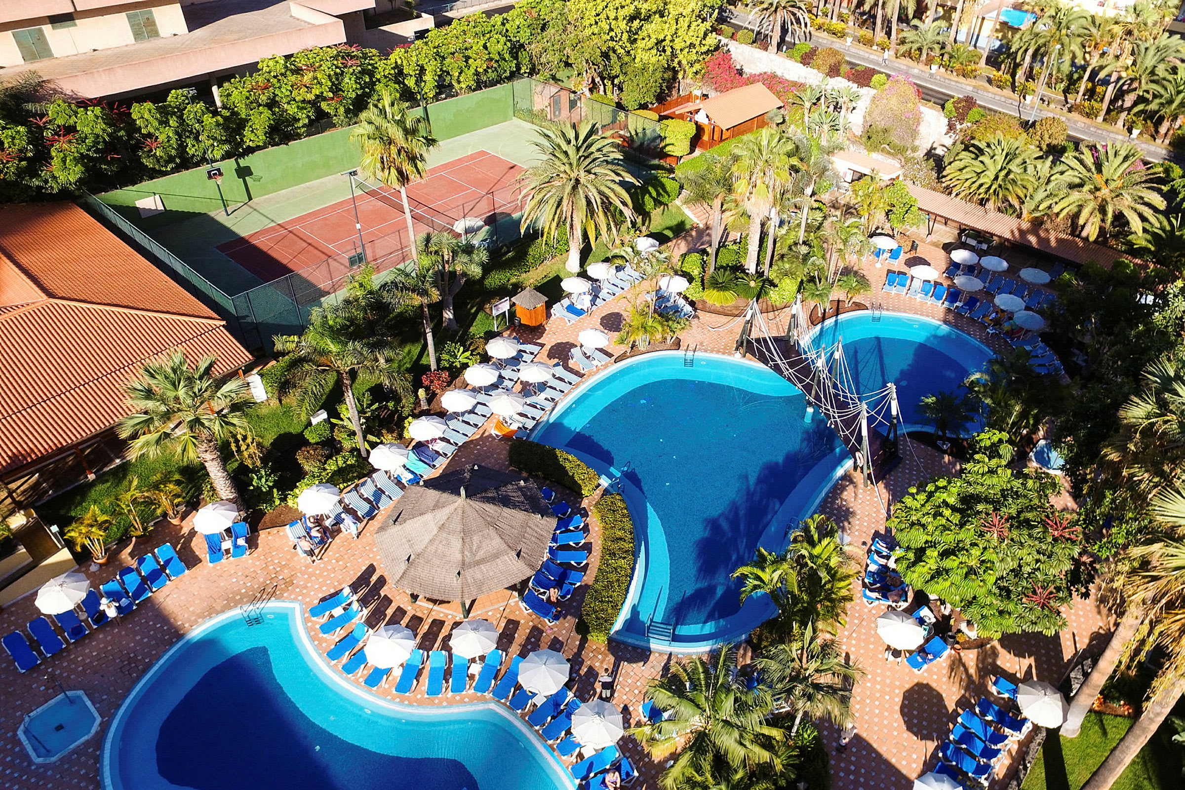 Vy över ett hotellområde med pooler, solstolar och parasoller i en grönskande miljö. (AI-genererad text)