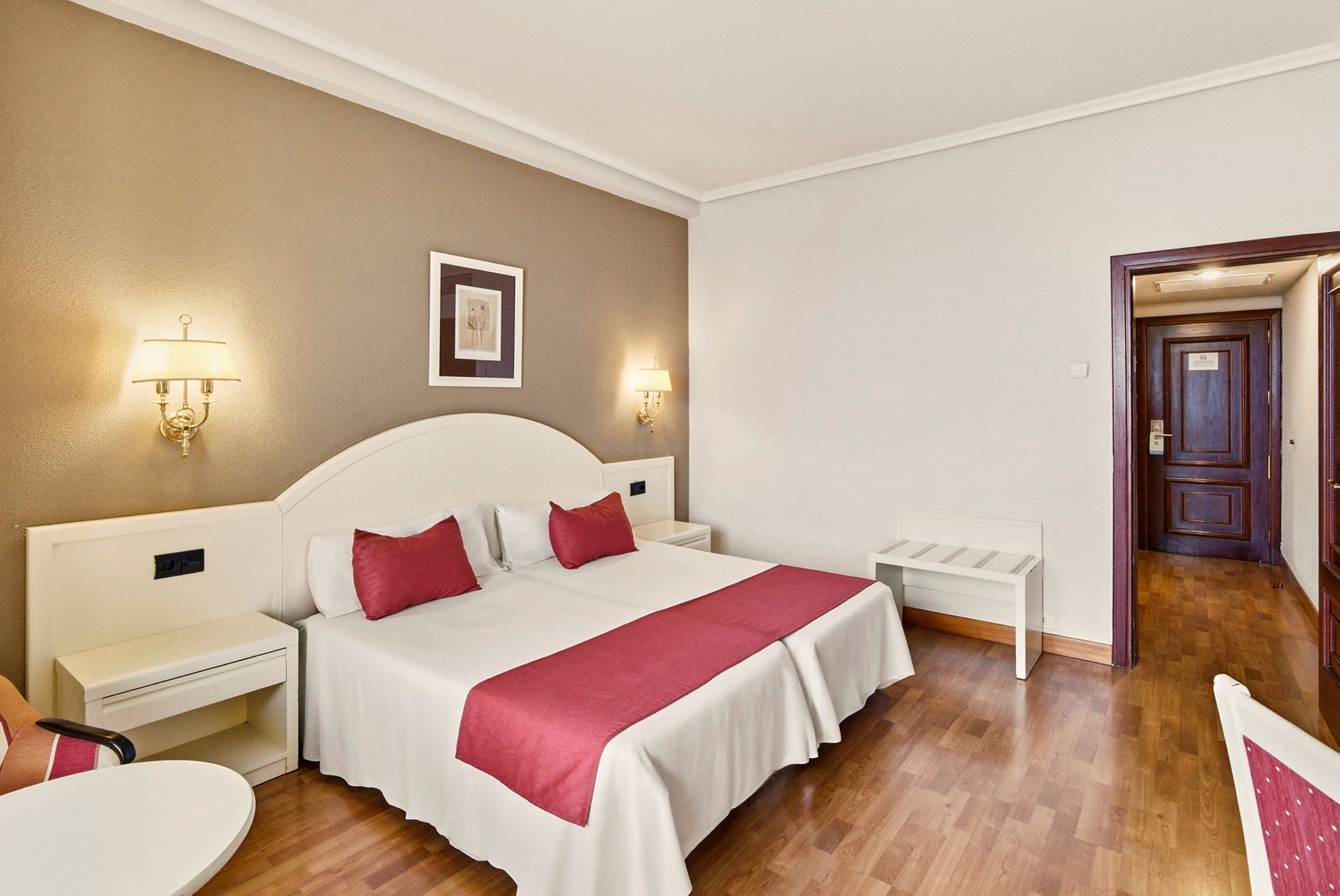 Ett hotellrum med en dubbelsäng som har röda detaljer, vägglampor och en dörr i bakgrunden. (AI-genererad text)