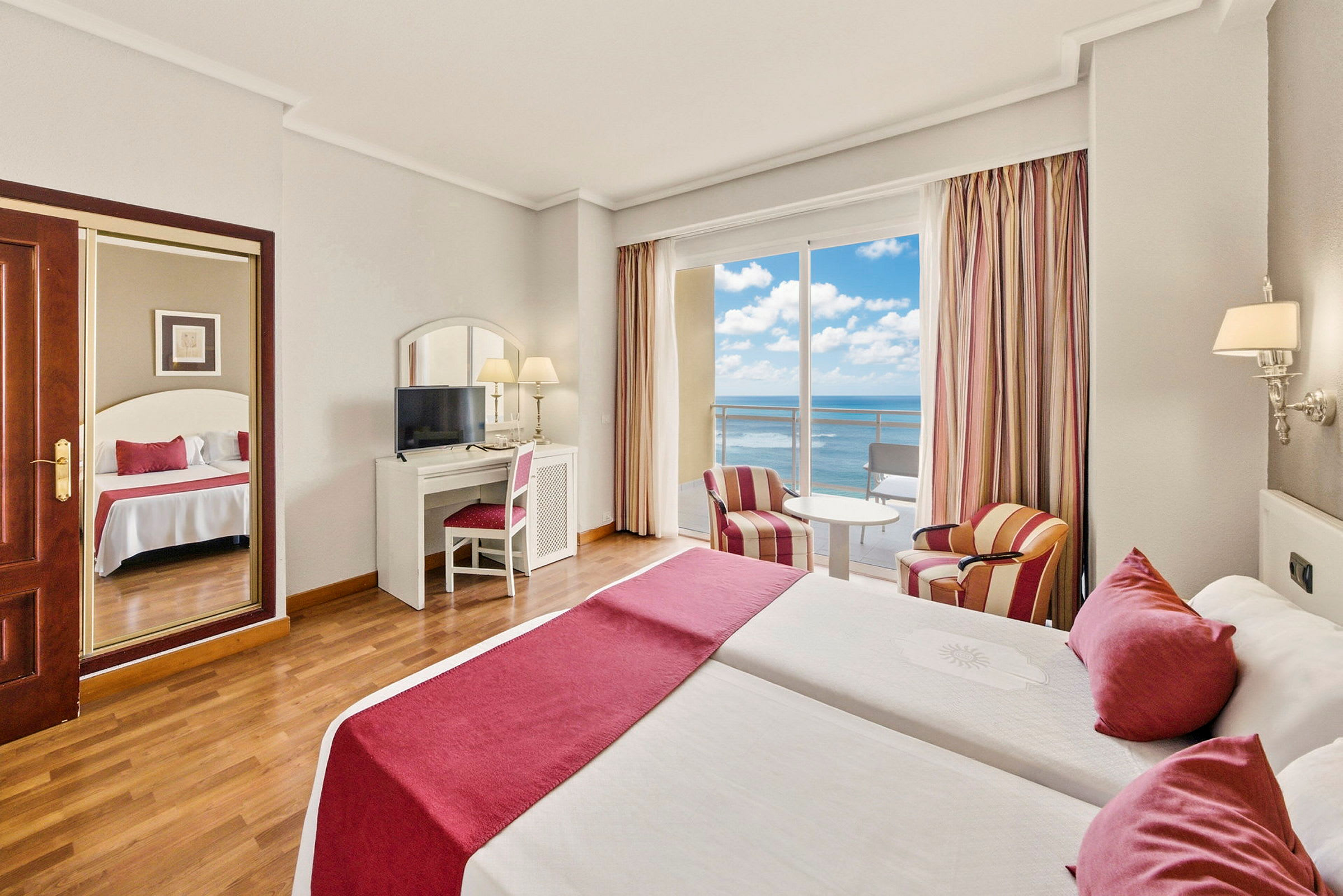 Hotellrum med dubbelsäng, sittmöbler, ett skrivbord med spegel och vy över havet genom stora fönster. (AI-genererad text)