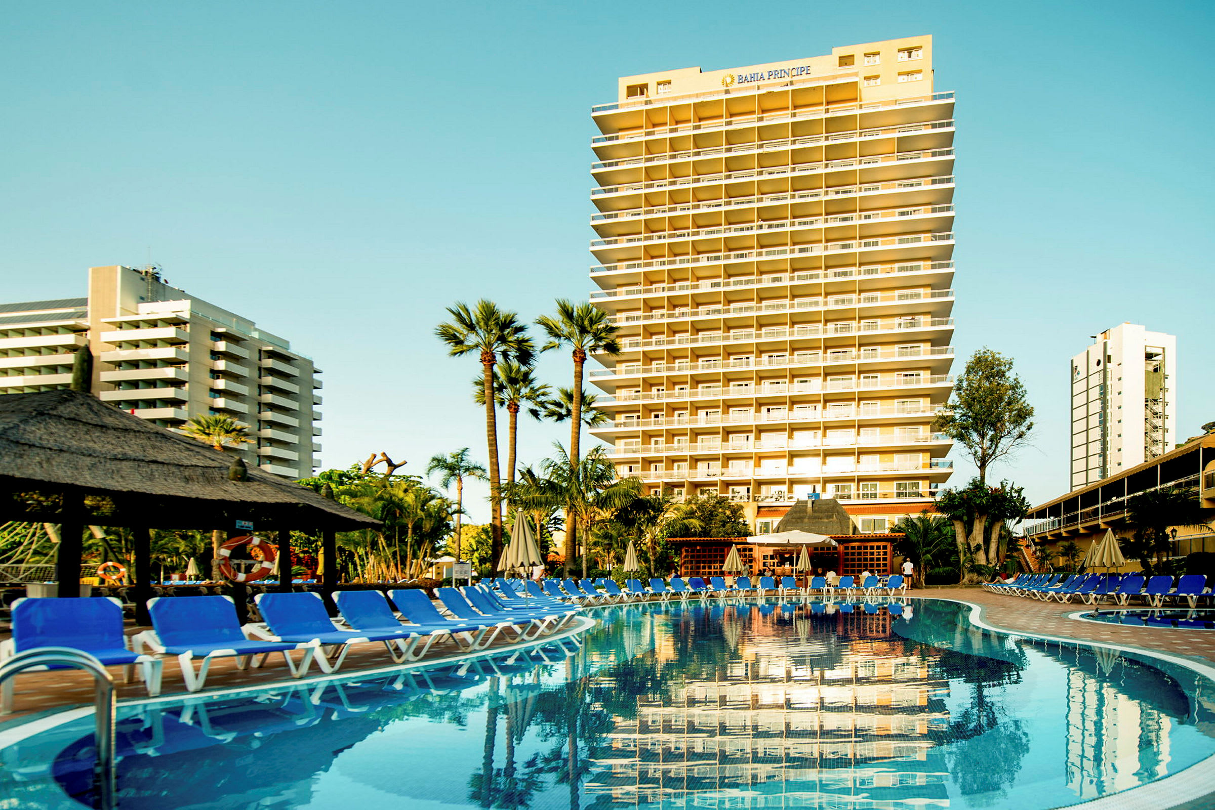 Utsikt över ett hotell med en stor pool och solstolar framför, omgiven av palmer. (AI-genererad text)