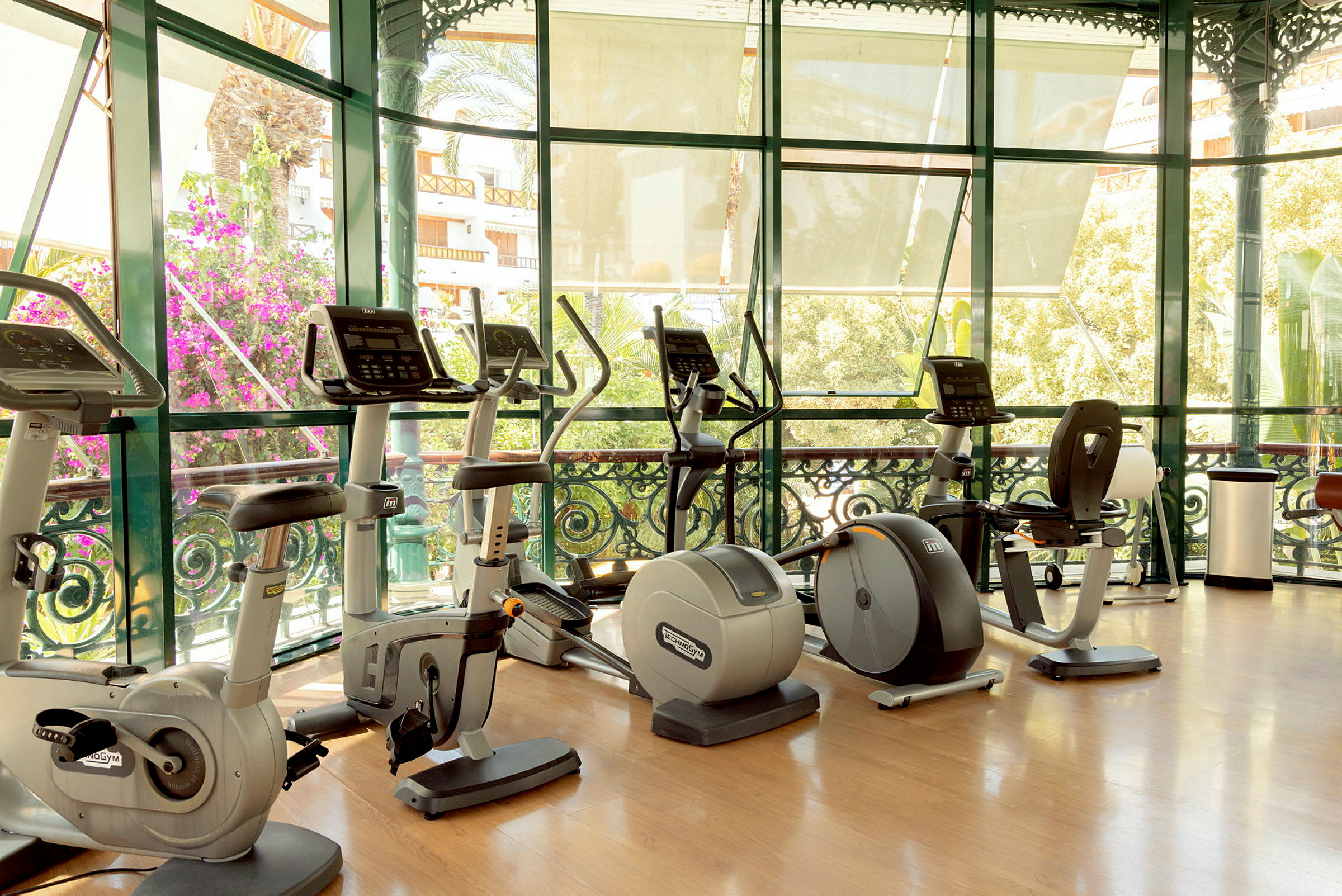Ett gym med träningscyklar och maskiner vid stora fönster med utsikt över träd och blommor. (AI-genererad text)