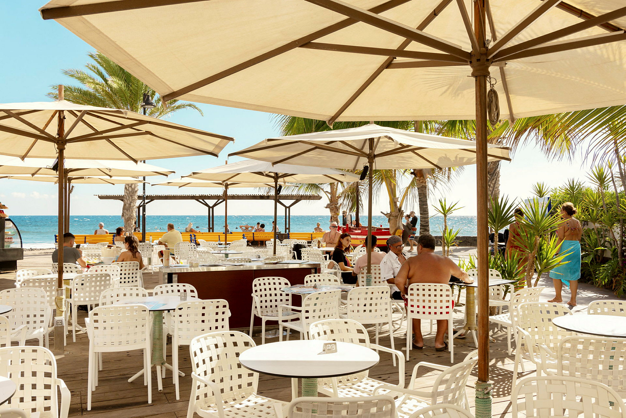 Utomhusrestaurang med vita bord och stolar under parasoller, utsikt mot havet. (AI-genererad text)
