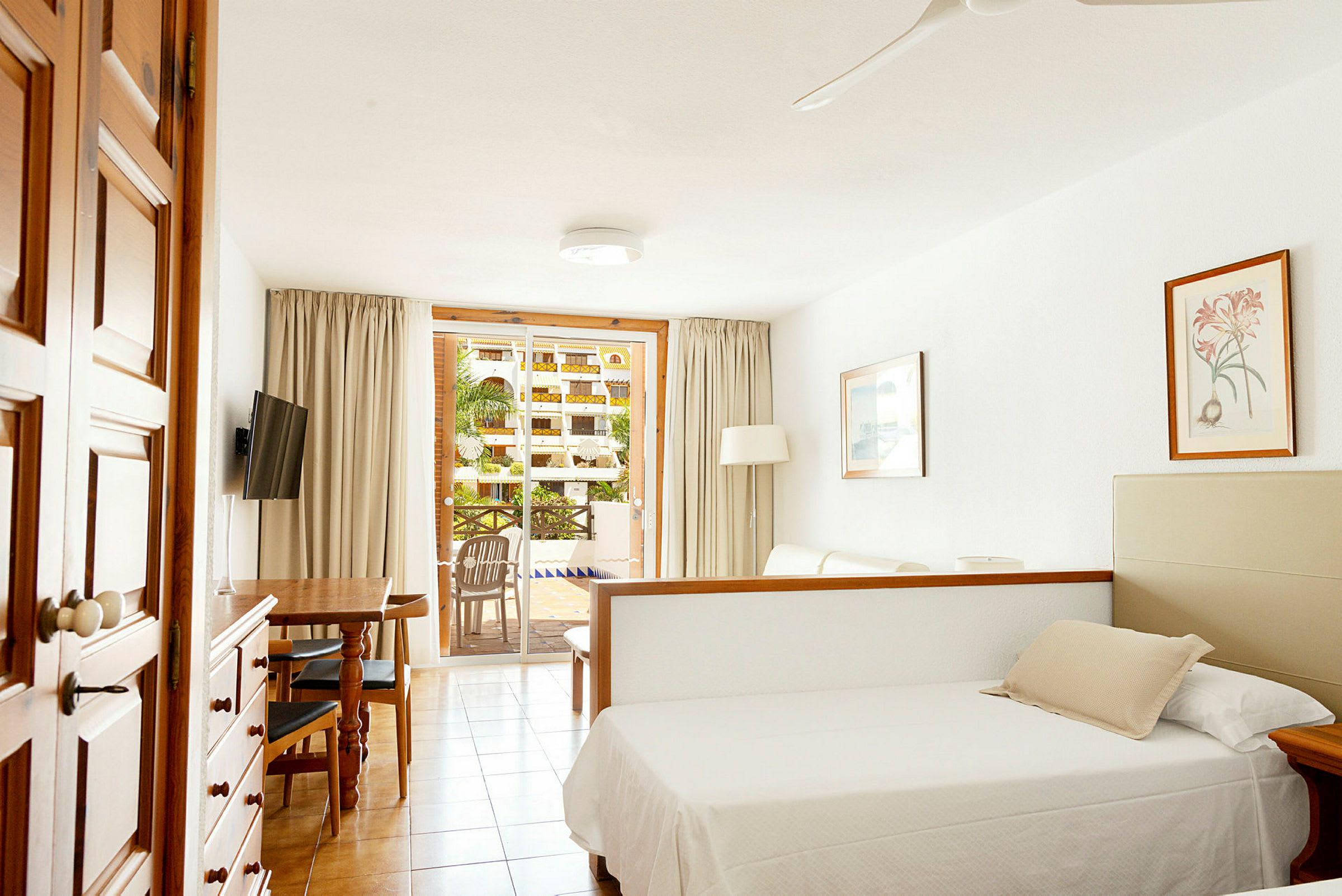 Ett hotellrum med en säng, soffa och matbord, samt utsikt mot en balkong med grönska utanför. (AI-genererad text)