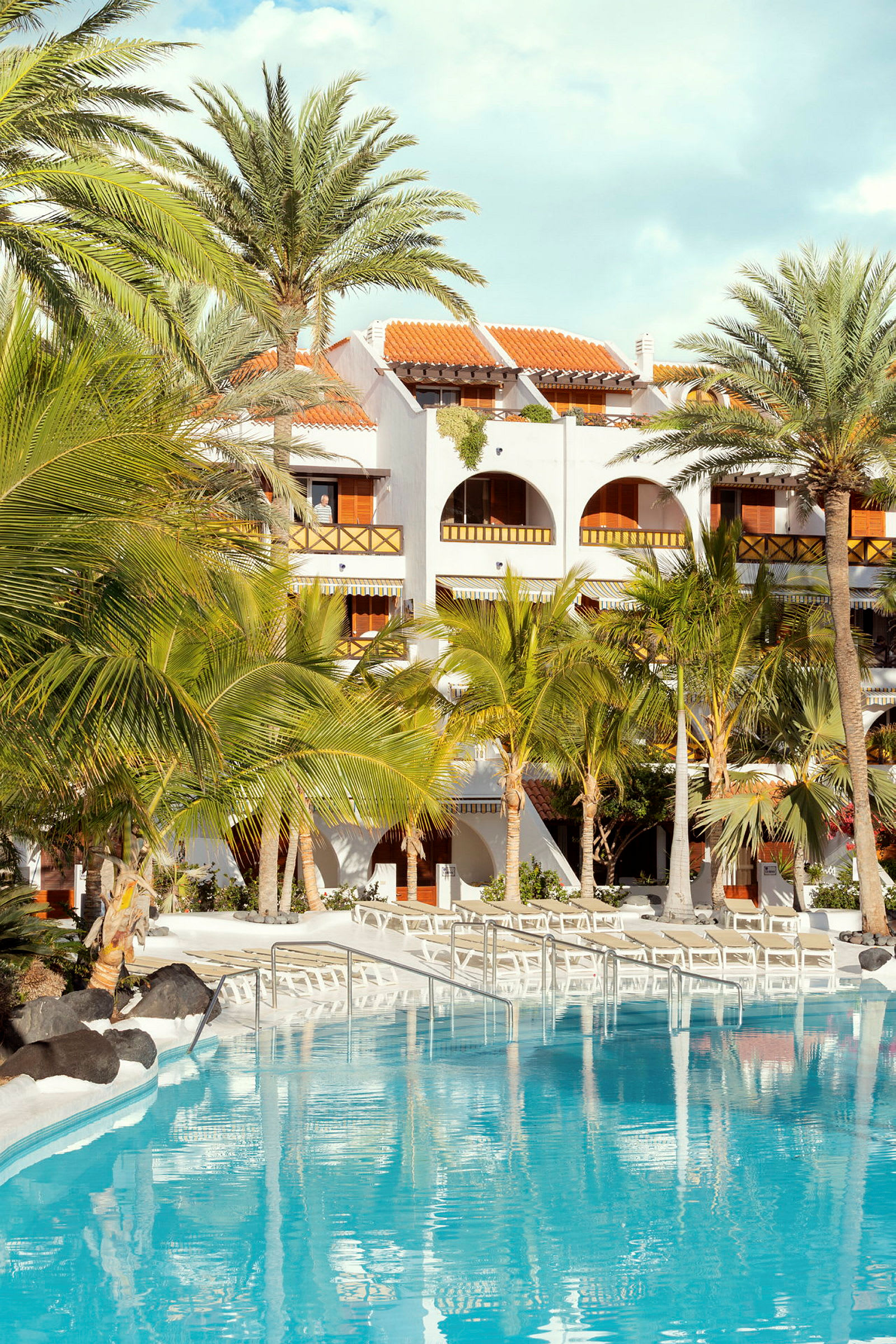 En hotellfasad med flera balkonger, omgiven av palmer, med en klar blå pool i förgrunden. (AI-genererad text)