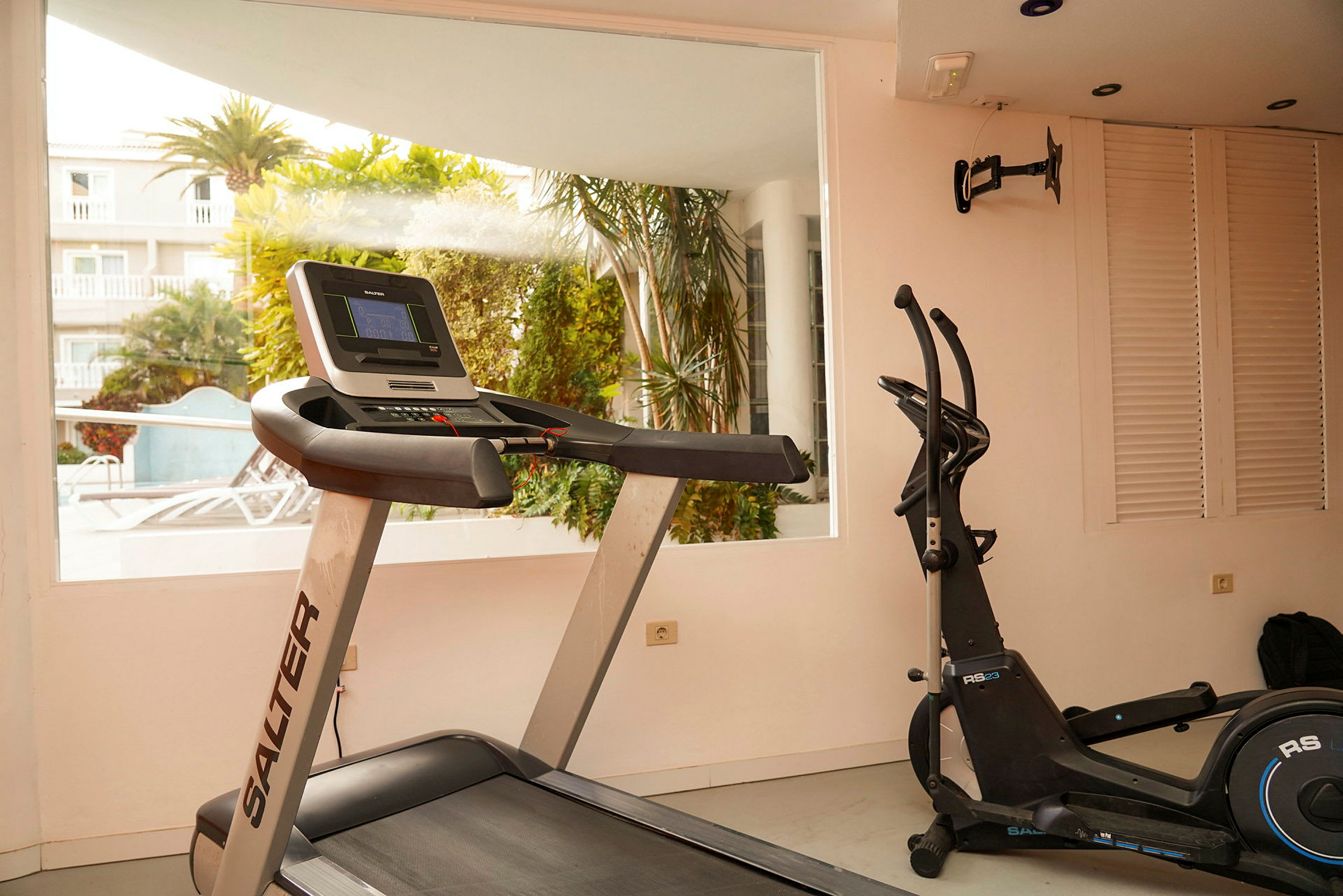 Ett gym med träningsutrustning, inklusive ett löpband och en motionscykel, med utsikt mot ett trädgårdsområde utanför. (AI-genererad text)
