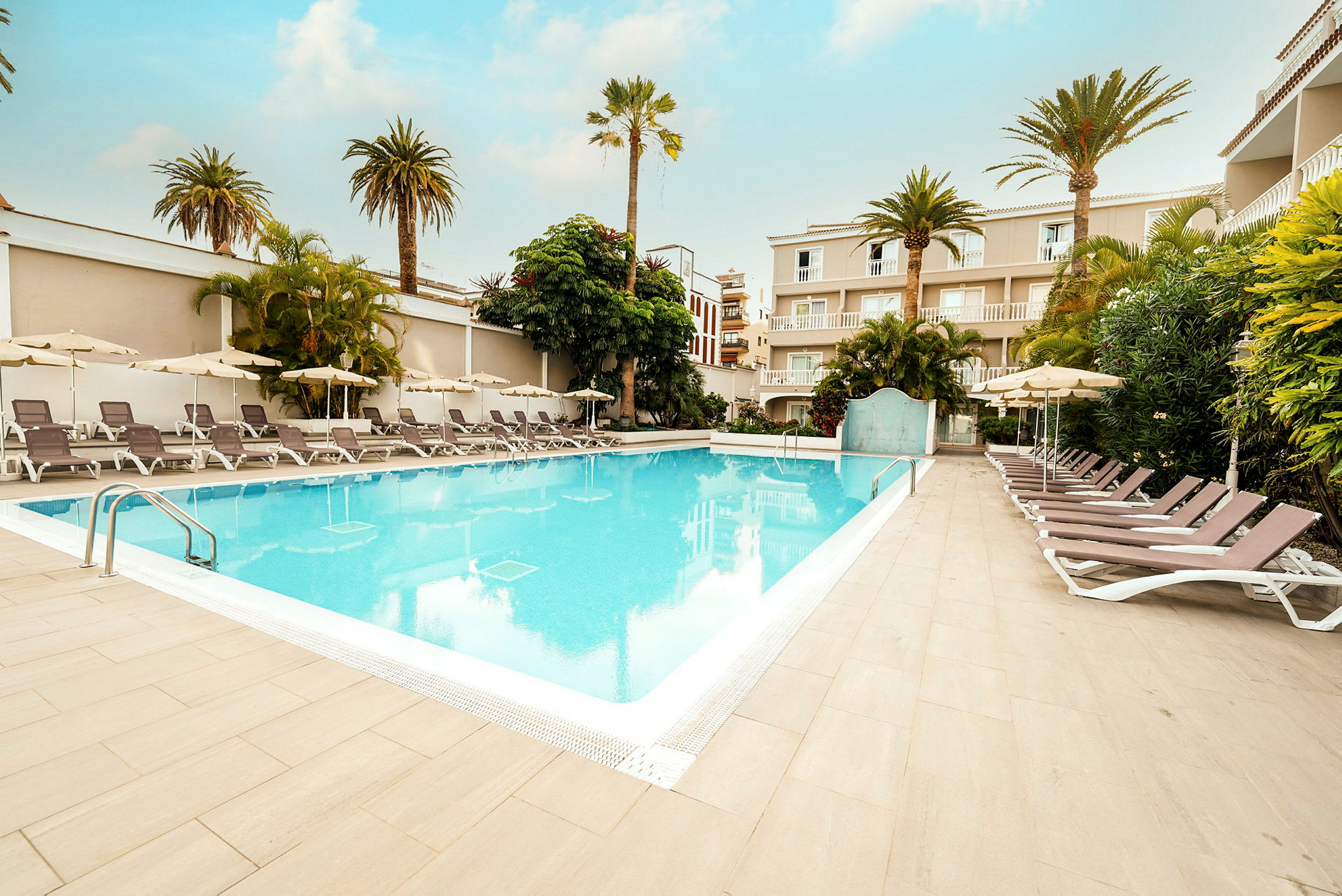 Utomhuspool med solstolar och parasoller omgiven av växtlighet och byggnader. (AI-genererad text)